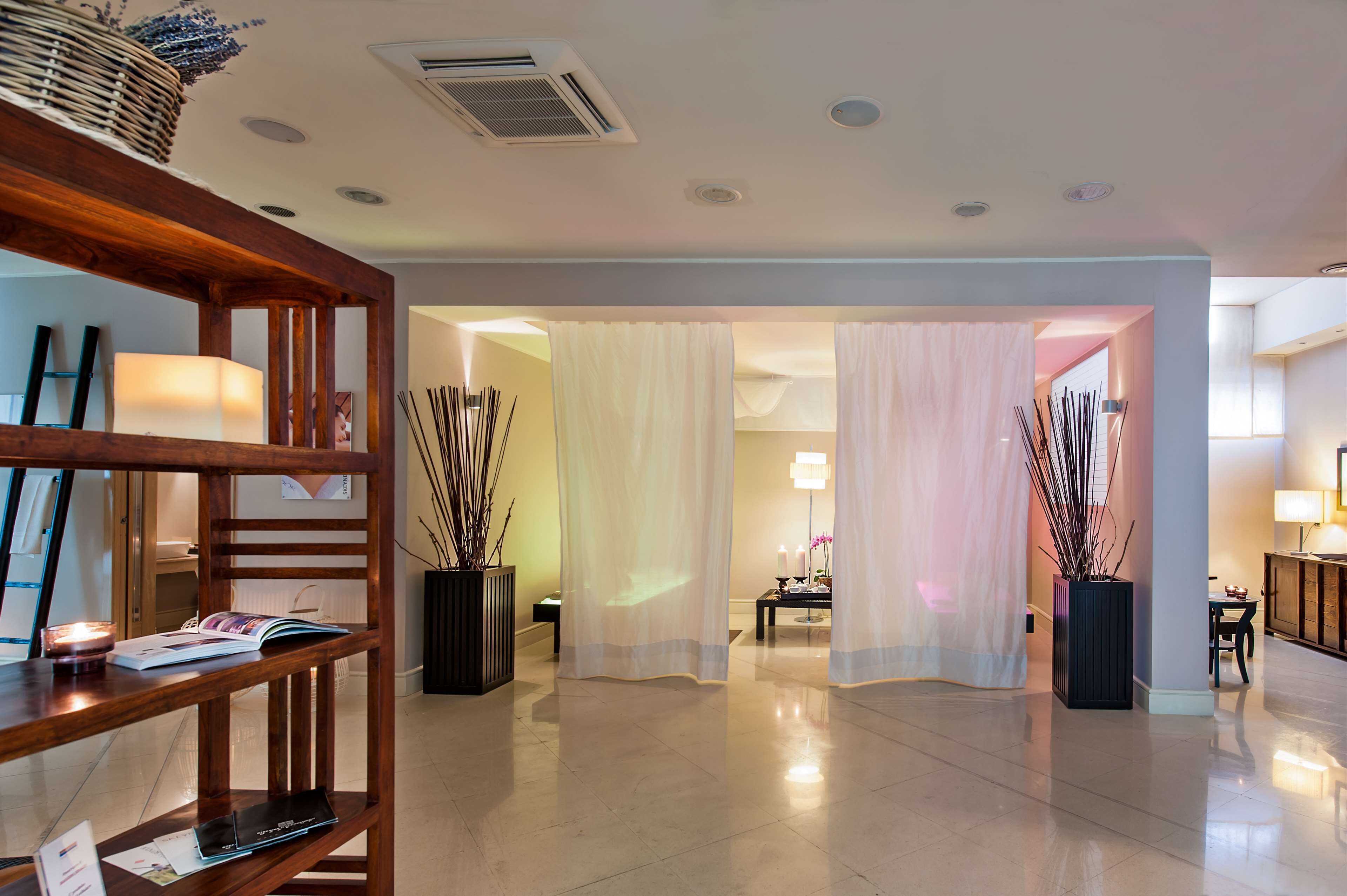 A modern hotel lobby with a wooden reception desk, a reading area with books and a lamp, and a view into a well-lit room through sheer curtains.
