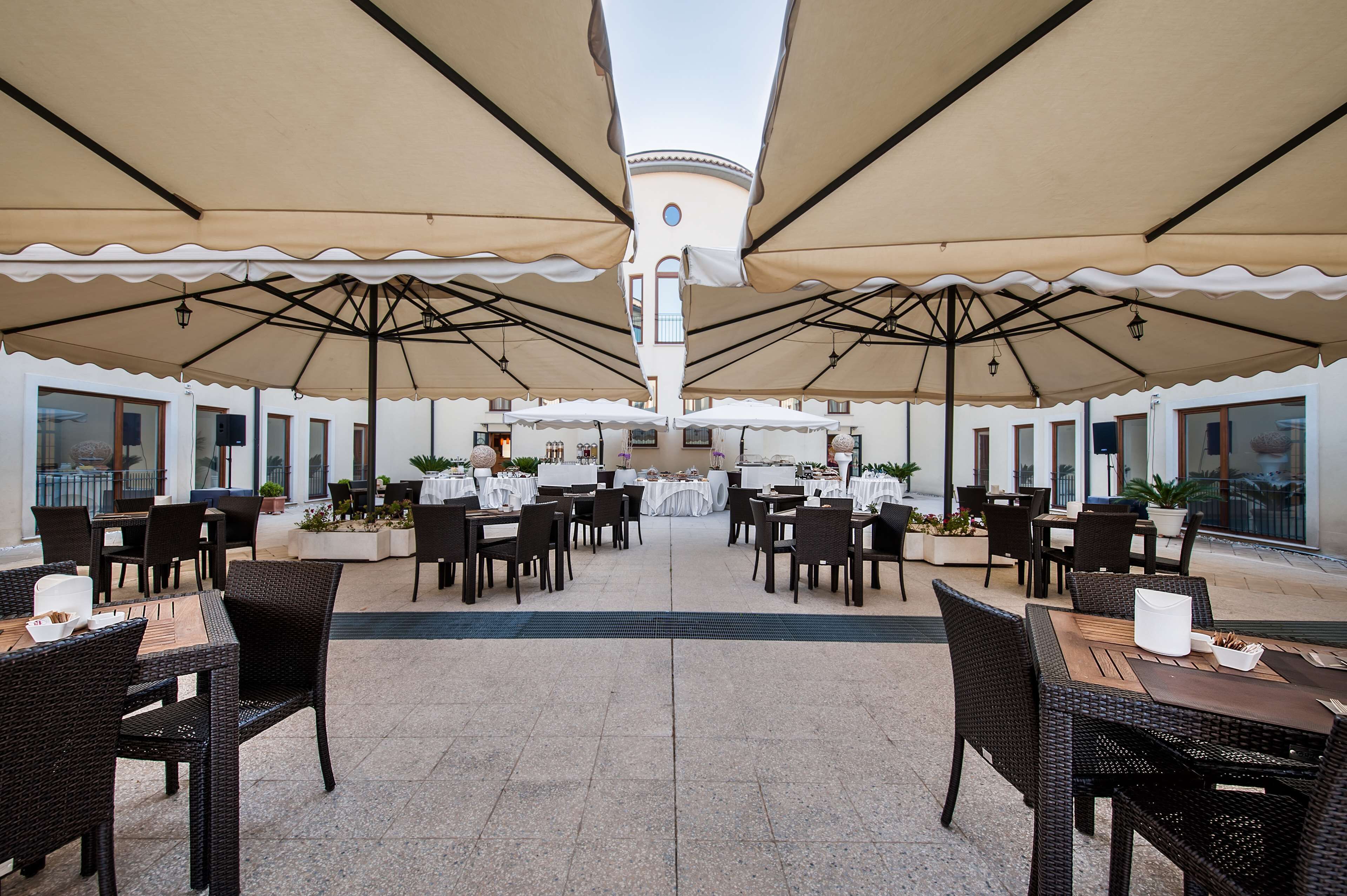Un área de comedor al aire libre con mesas y sillas, cubierta por grandes sombrillas, y una vista central de un edificio.