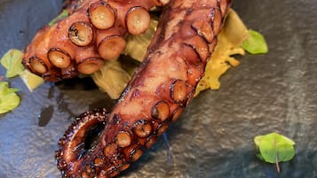Ein Detailaufnahme von gegrilltem Oktopus mit gebräunter Haut und Tentakeln, serviert auf einer dunklen Oberfläche mit grünen Blättern.