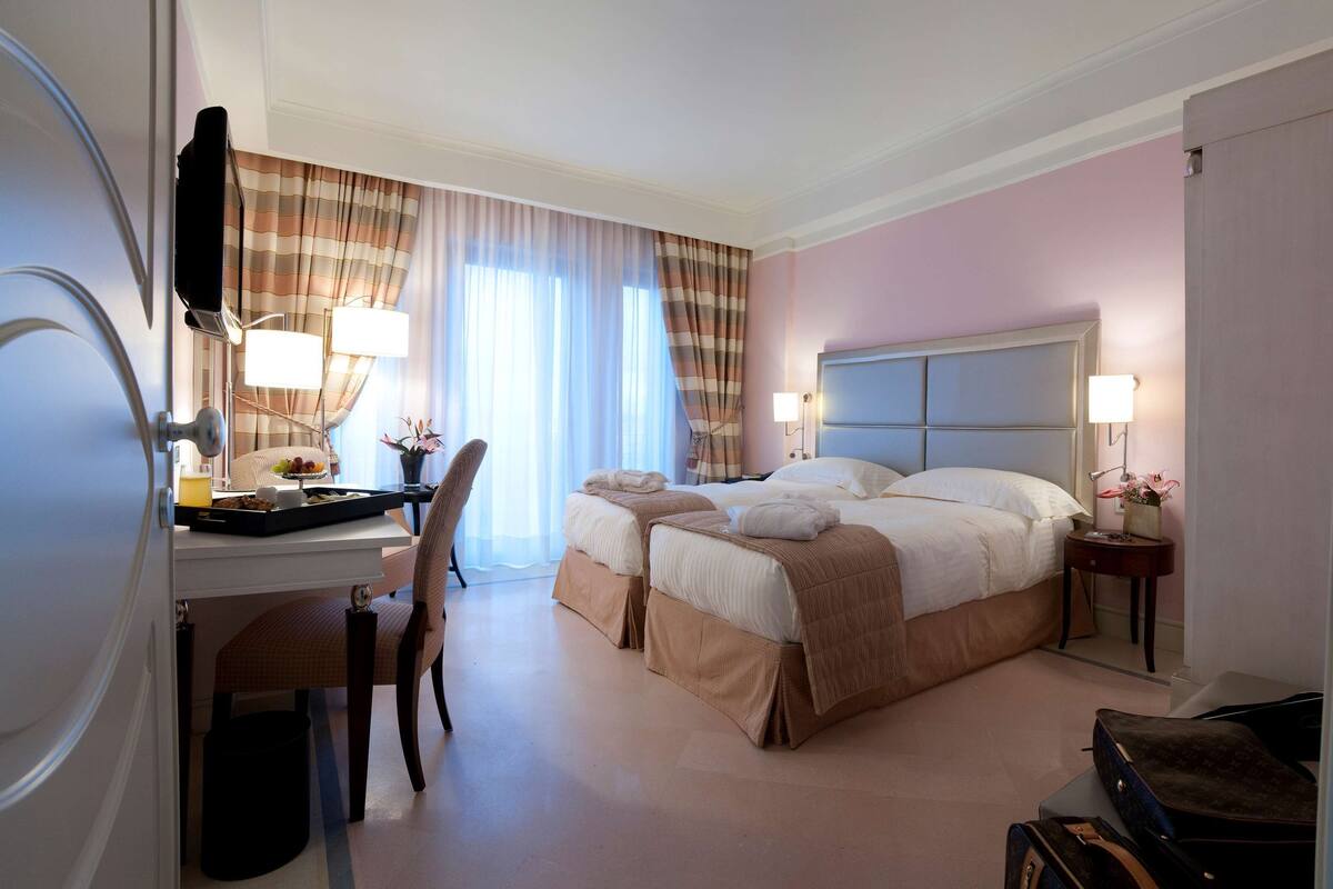 A hotel room with a bed, two bedside tables, a desk with a chair, a lamp, and a window with curtains.
