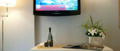 A hotel room with a flat-screen TV mounted on the wall, a white console table with a bottle and glasses, and a potted plant.