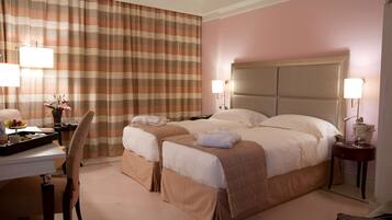 A hotel room with two beds, a desk, and a chair. There are lamps on the nightstands and a tray with fruits on the desk.