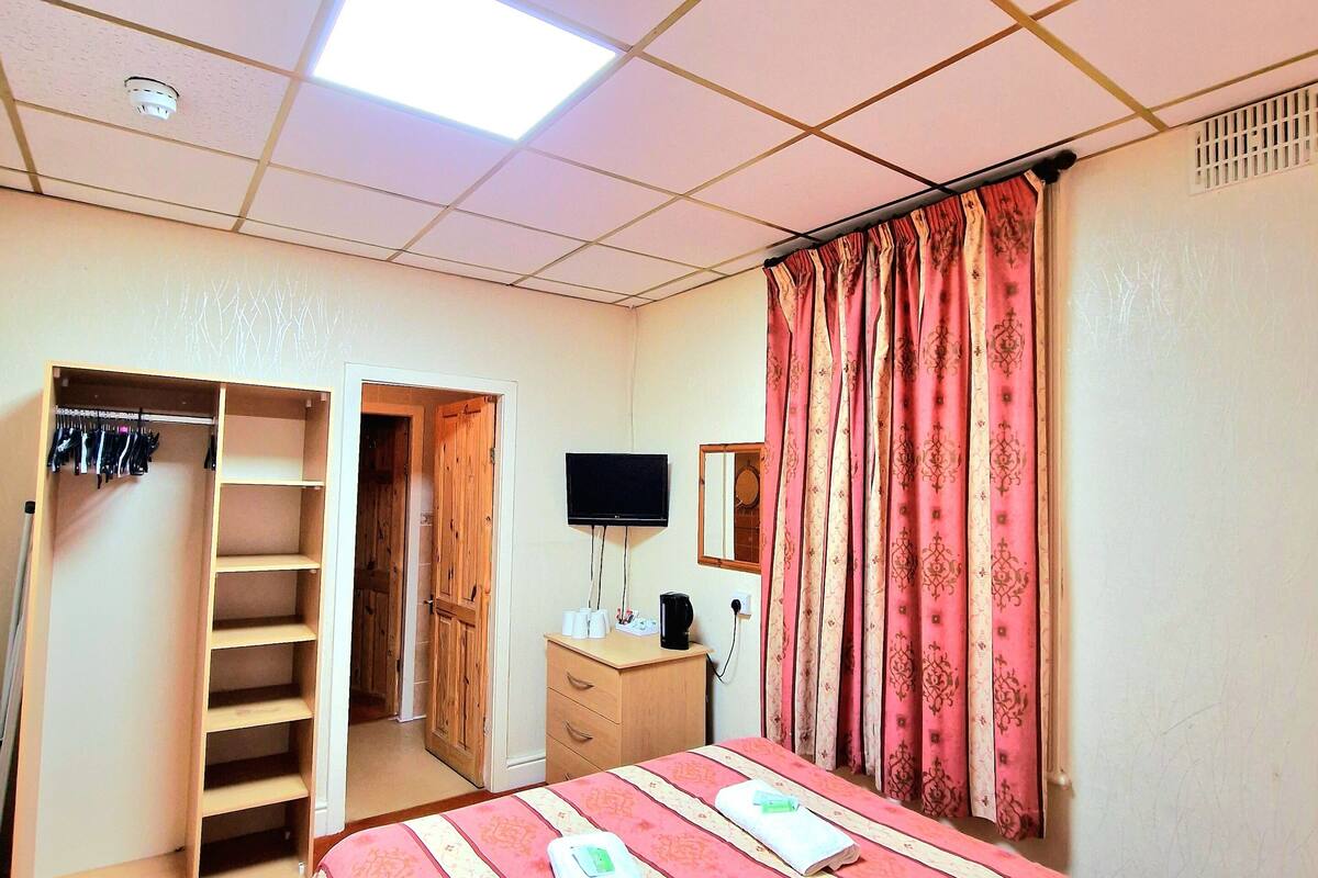 A bedroom with a bed, a wardrobe, a desk with a flat-screen TV, and a window with curtains.
