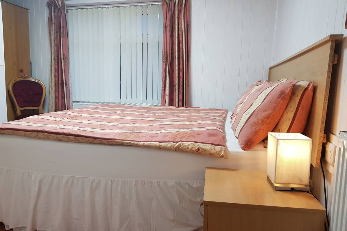 A bedroom with a bed, a bedside table with a lamp, a chair, and a window with curtains.