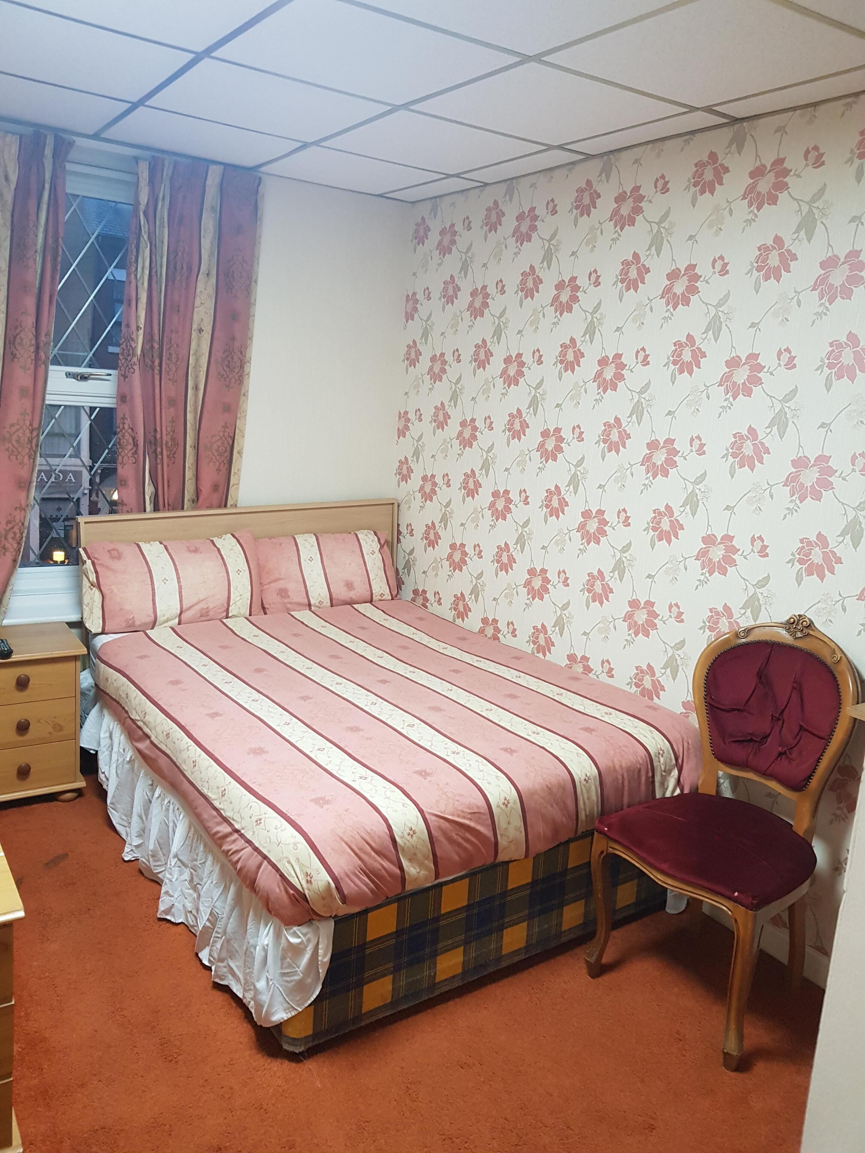 A bedroom with a bed, a wooden nightstand, a chair, and a window with curtains.