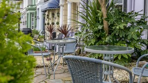 An outdoor seating area with wicker chairs and round tables, surrounded by greenery and a building with a blue awning. - The Belmont (Torquay)