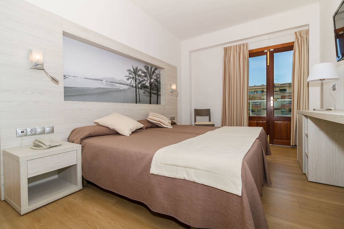 Habitación de hotel con una cama grande, una mesita de noche con un teléfono, un cuadro de una playa y vistas al exterior a través de una puerta.
