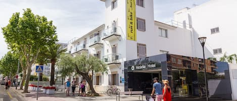 A hotel with a sign reading "Alcudia Hotel" and a pedestrian area with people and a stroller.