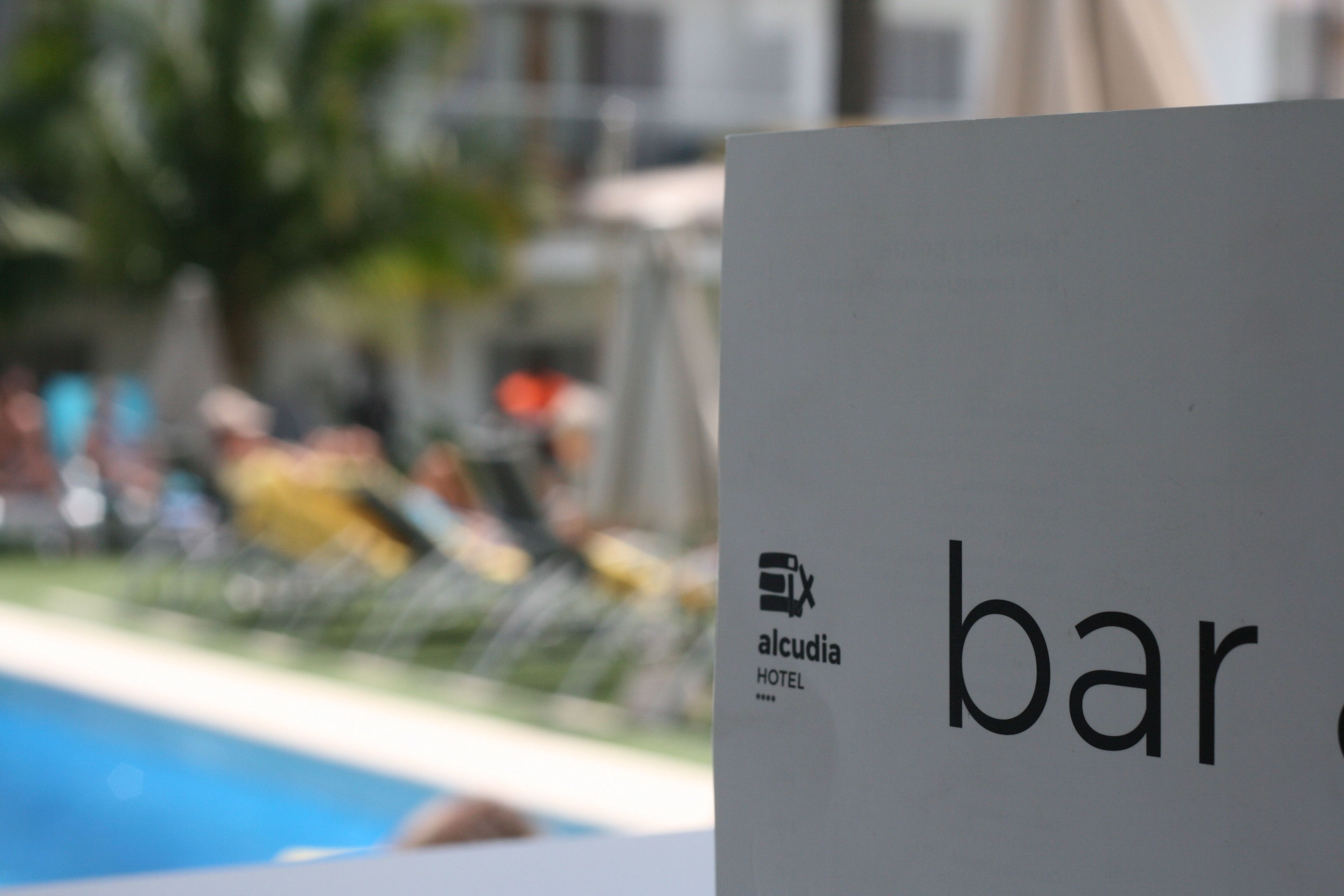 Skilt for Alcudia Hotel med delvis udsigt til en pool og en uskarp baggrund.