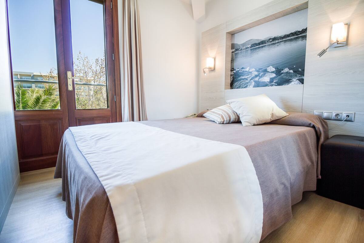 Un dormitorio con cama, mesita de noche, lámpara de pared y ventana con cortinas.