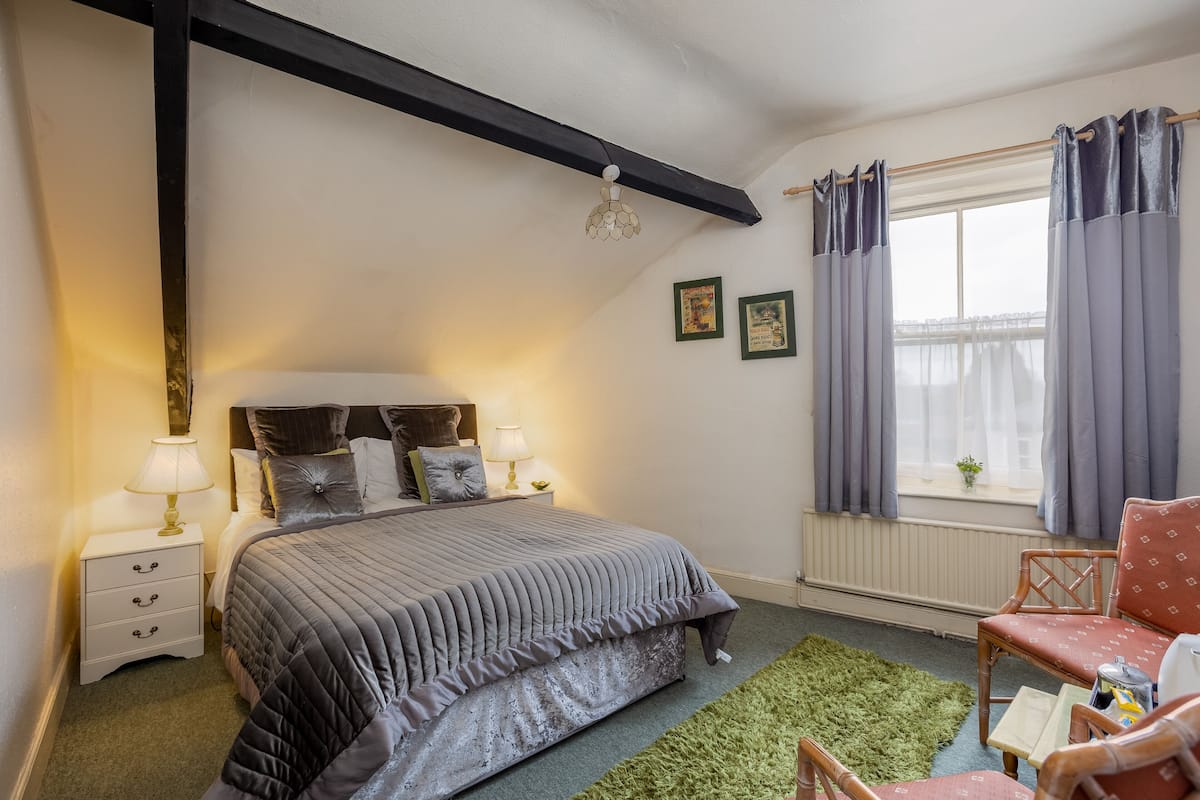 A bedroom with a bed, bedside tables, a chair, a window with curtains, and a radiator.