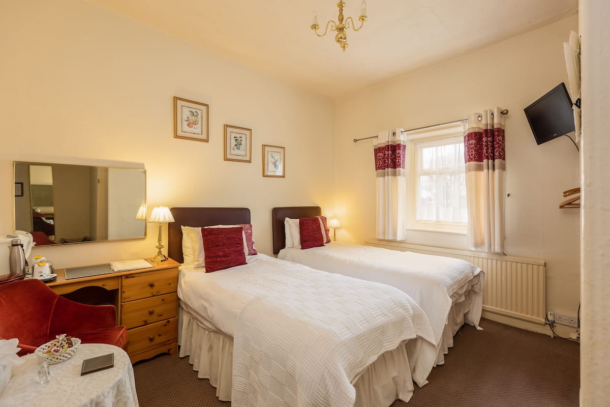 A hotel room with two beds, a desk with a lamp, a chair, a small table, a mirror, and a TV on the wall.
