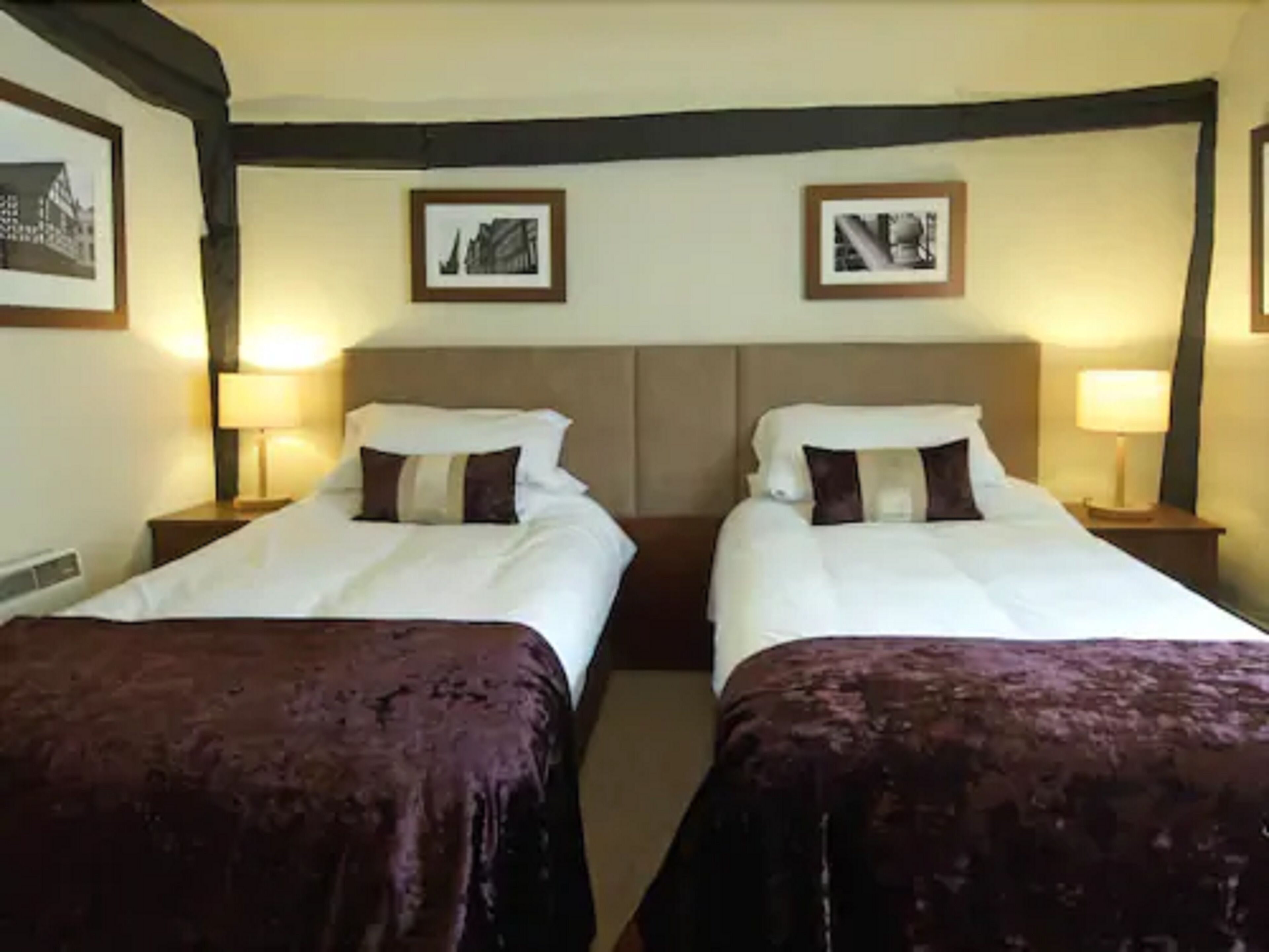 A hotel room with two single beds, each with a brown bedspread and white linens. There are two bedside tables with lamps, and the walls are adorned with framed pictures.