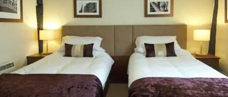 A hotel room with two single beds, each with a brown bedspread and white linens. There are two bedside tables with lamps, and the walls are adorned with framed pictures.