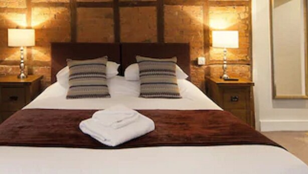 A neatly made bed with a brown blanket and two pillows, flanked by two bedside tables with lamps, in a room with exposed brick walls. - The Talbot Inn (Ledbury)