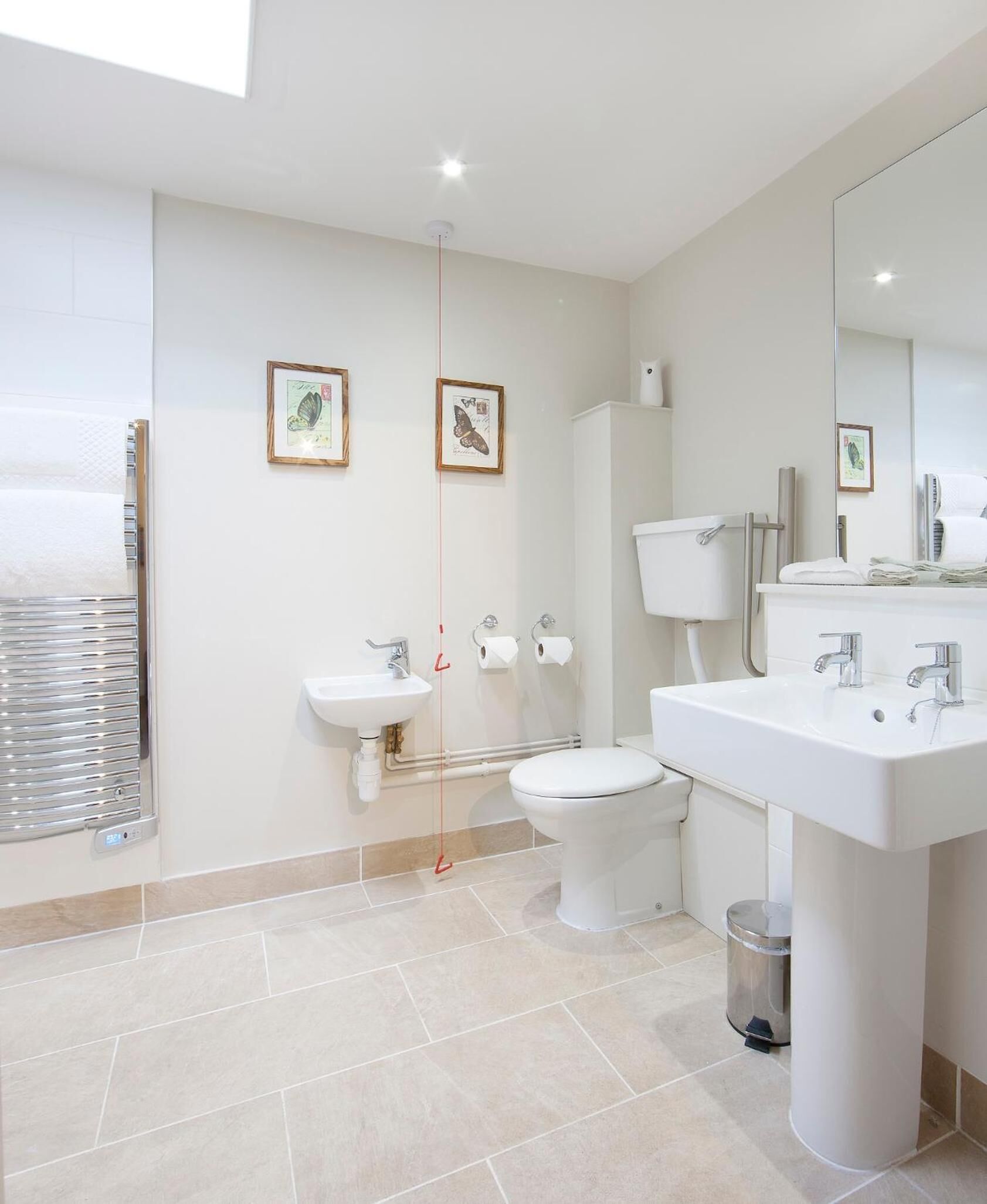 A modern bathroom with a toilet, bidet, and sink. There are framed pictures on the wall, a window with blinds, and a trash can.