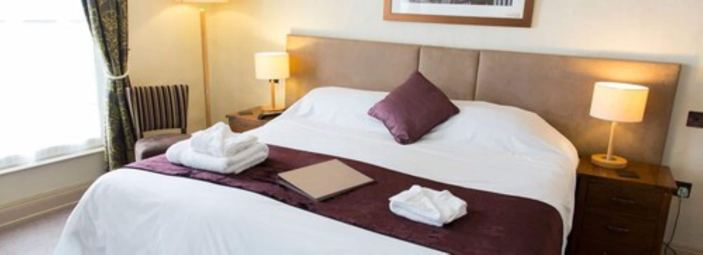 A neatly made bed with white and purple bedding, a folded towel, and a book on the bed. There are two bedside lamps and a nightstand with a lamp.