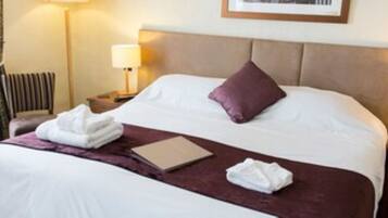 A neatly made bed with white and purple bedding, a folded towel, and a book on the bed. There are two bedside lamps and a nightstand with a lamp.