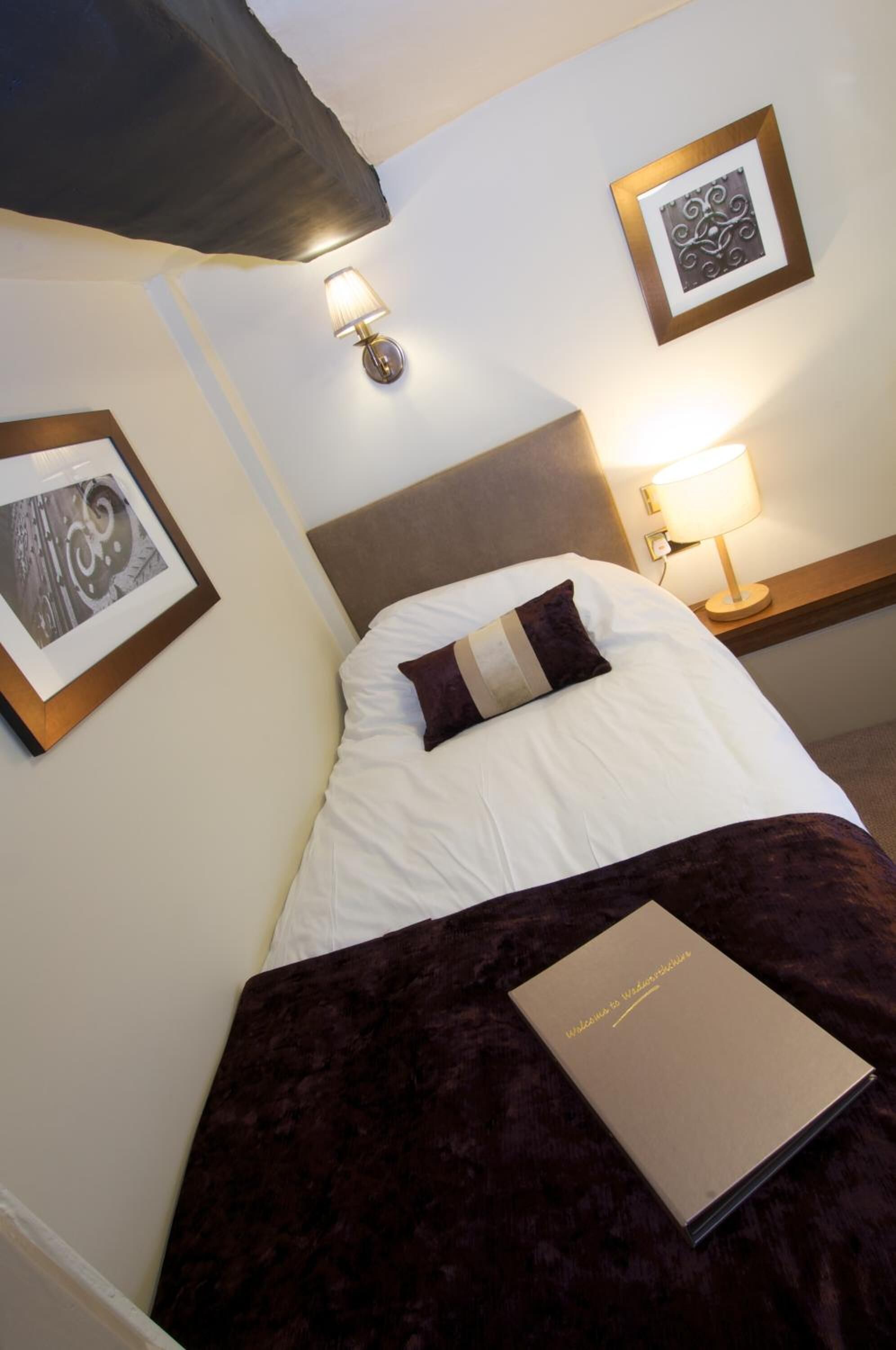 A neatly made bed with a dark blanket and a single pillow, a framed picture on the wall, a bedside lamp, and a book on the bed.