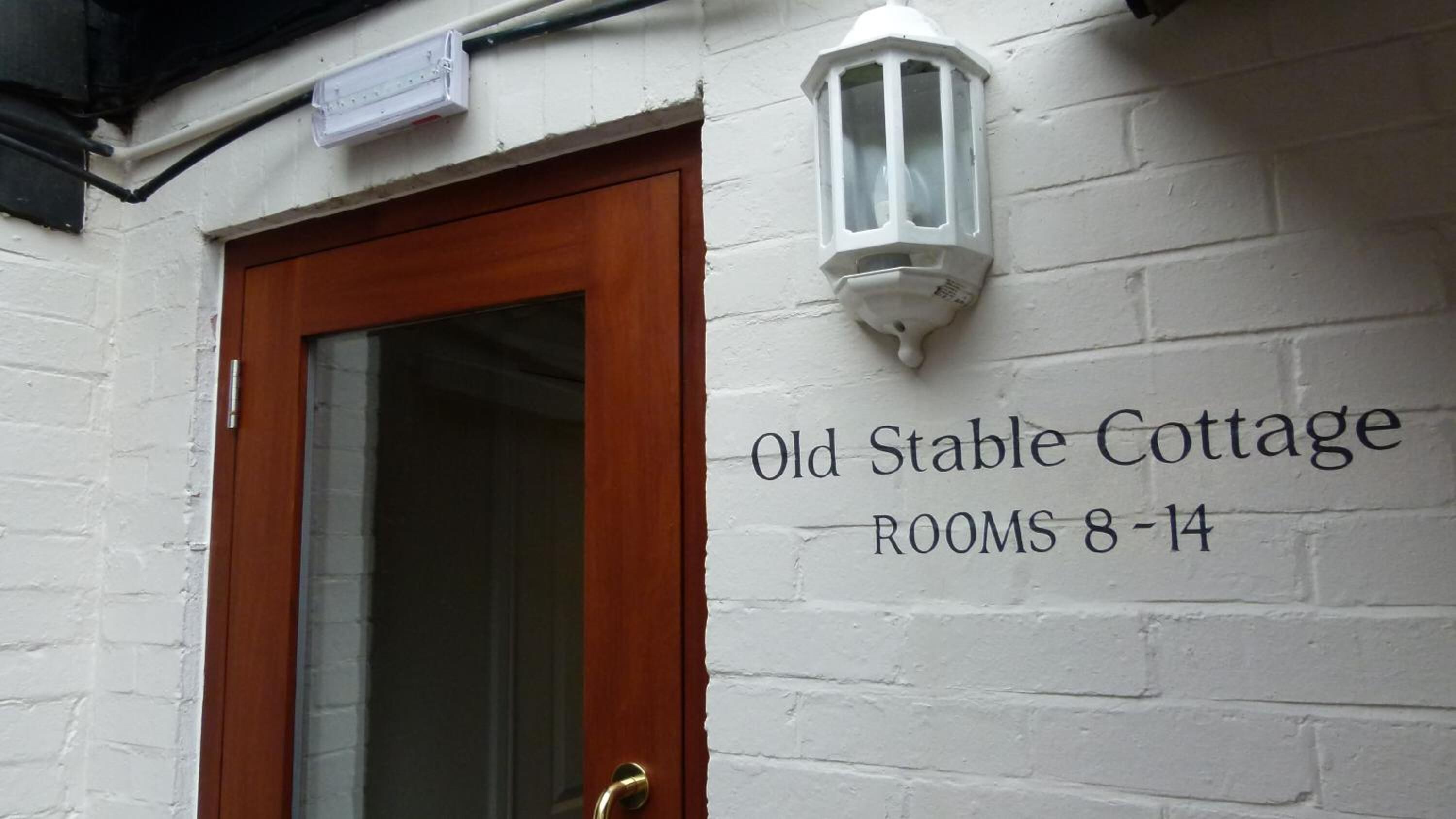 A white brick wall with a wooden door and a sign reading "Old Stable Cottage ROOMS 8-14".