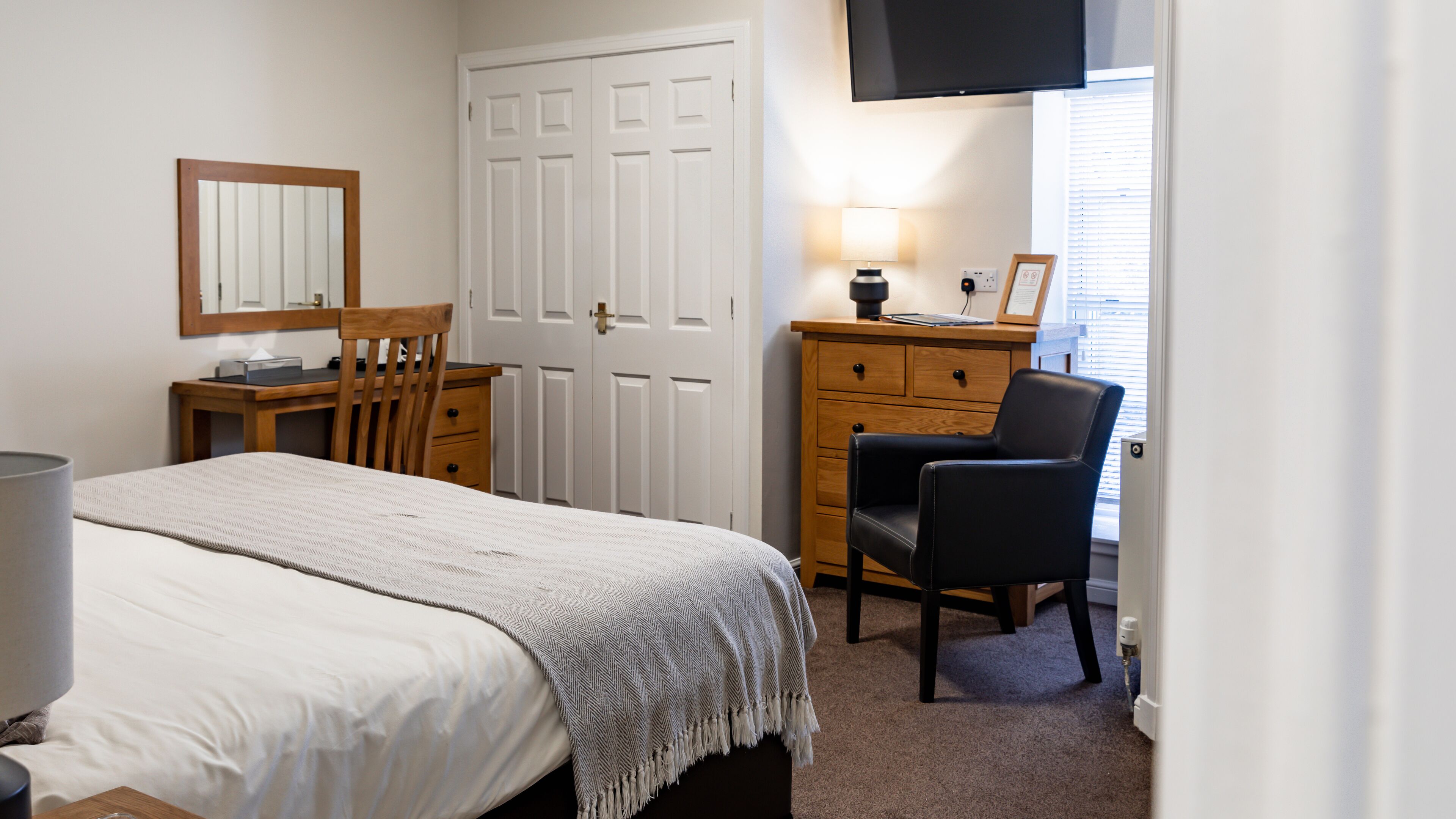 A bedroom with a bed, a wooden desk, a chair, a dresser, a mirror, and a television.