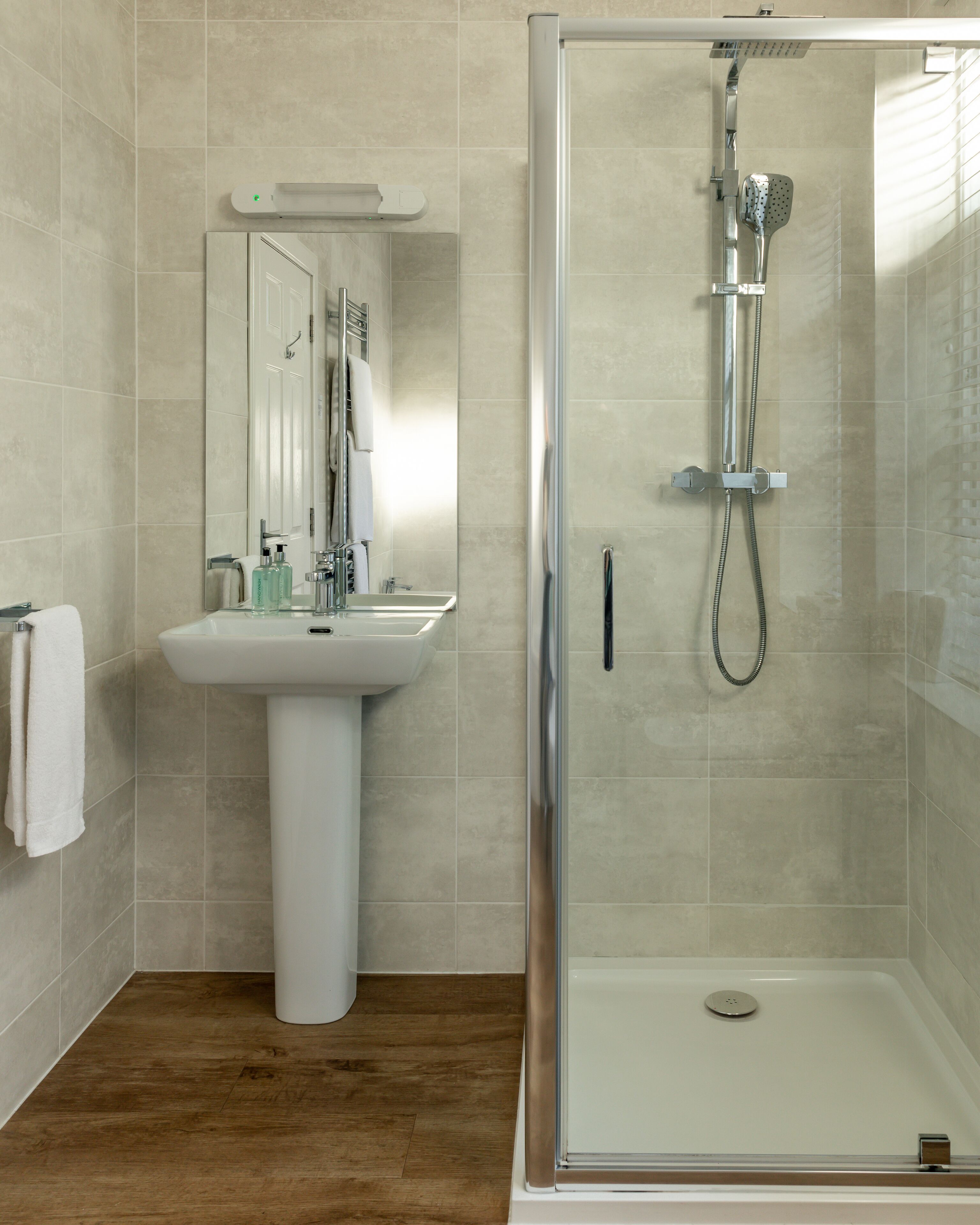 A modern bathroom with a pedestal sink, a glass-enclosed shower, and a wooden floor.