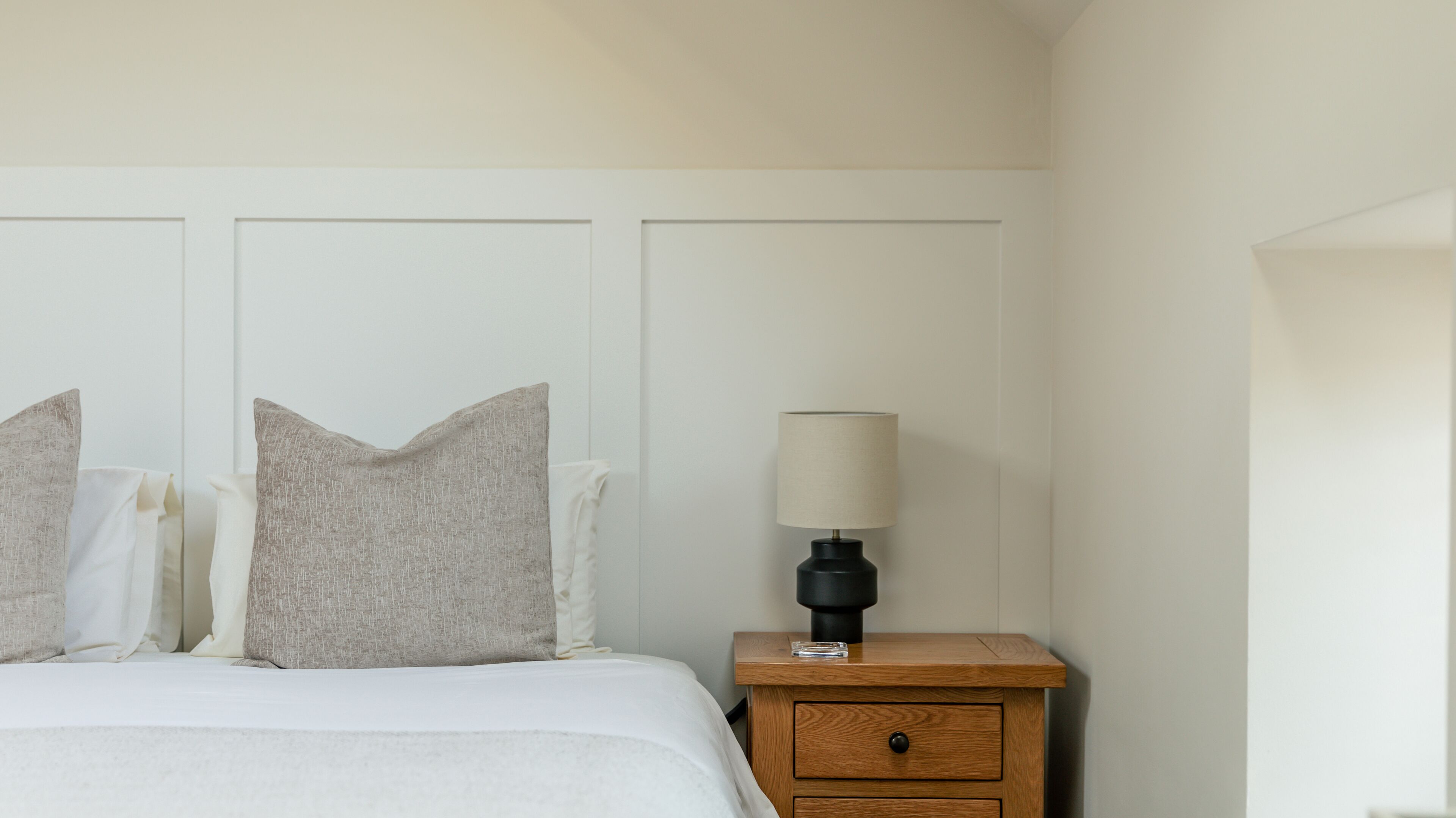 A neatly made bed with white linens and two pillows, a bedside table with a lamp, and a wooden nightstand with a drawer.