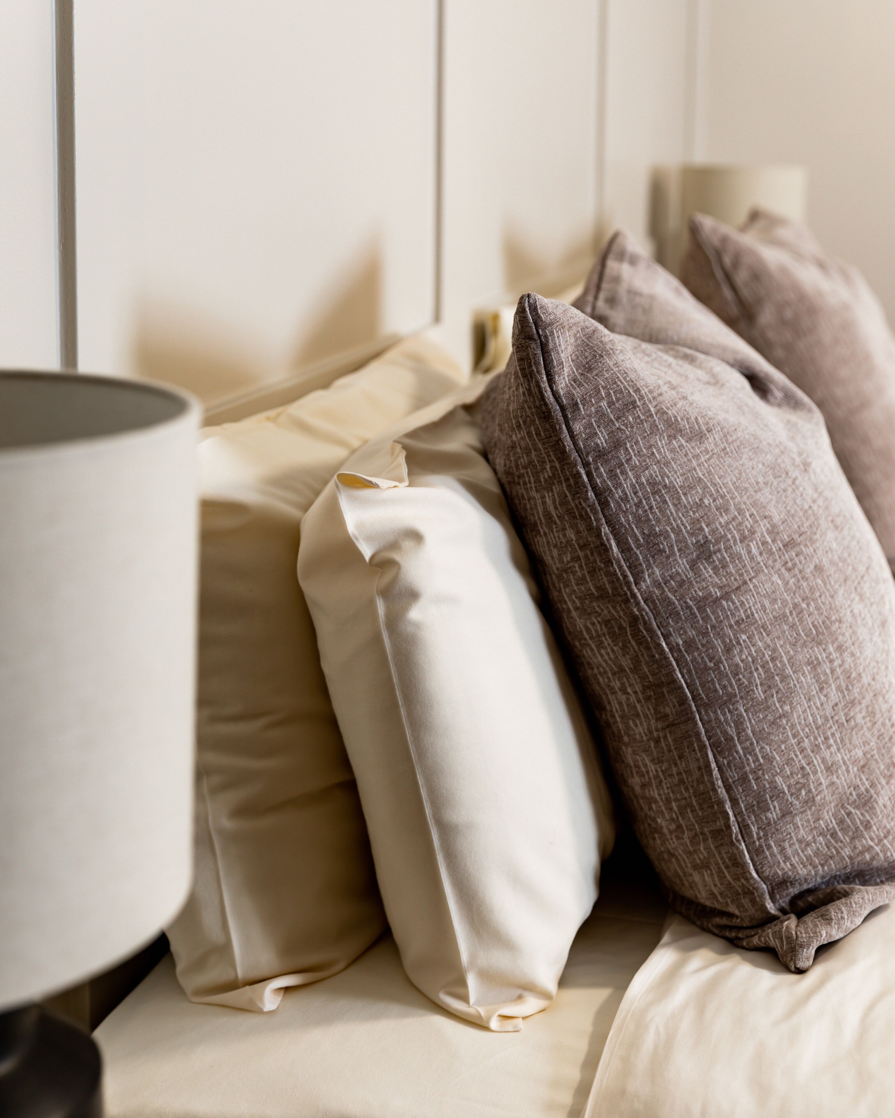 A bed with beige and gray pillows, a white headboard, and a bedside lamp.