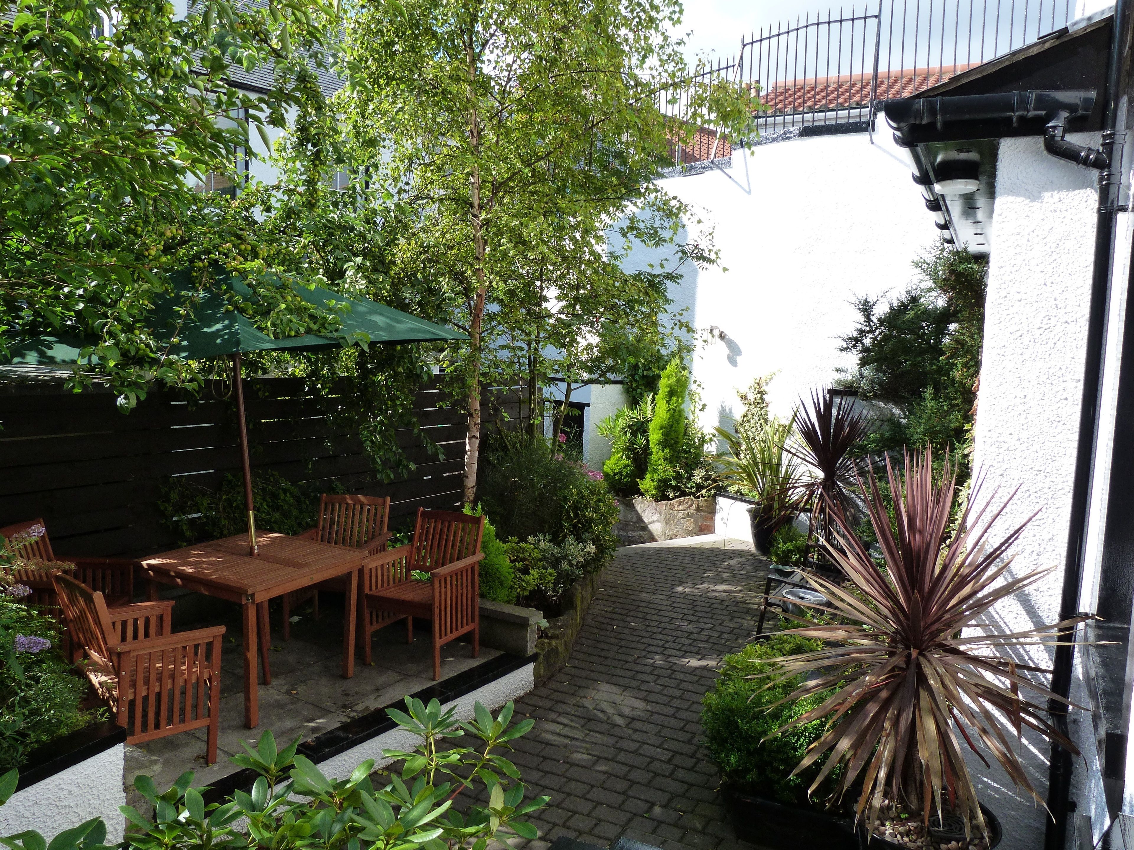 Un patio con mesa y sillas de madera, rodeado de plantas en macetas y un paraguas verde.