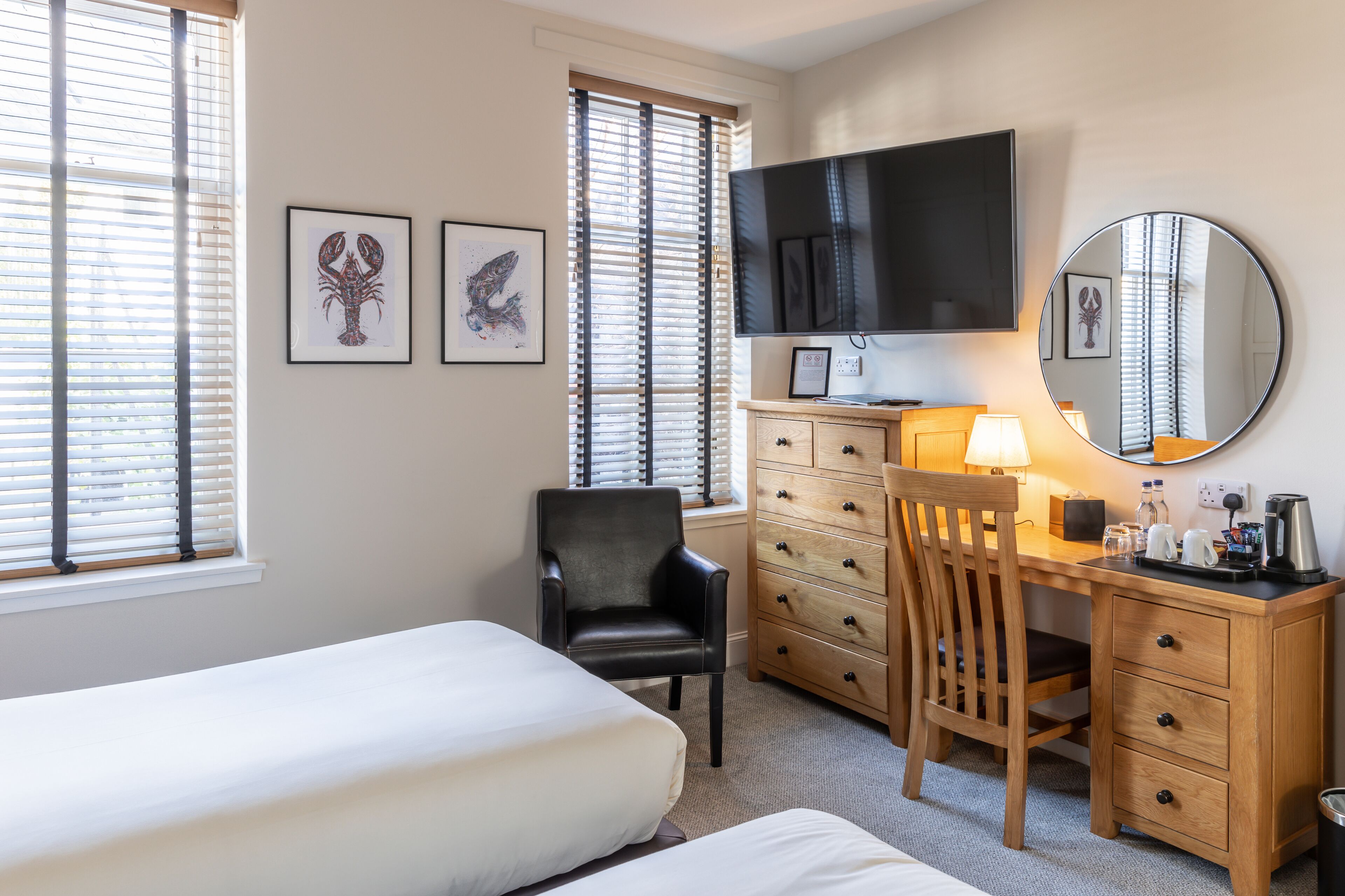 A hotel room with a bed, a desk with a chair, a TV, a mirror, and artwork on the wall.