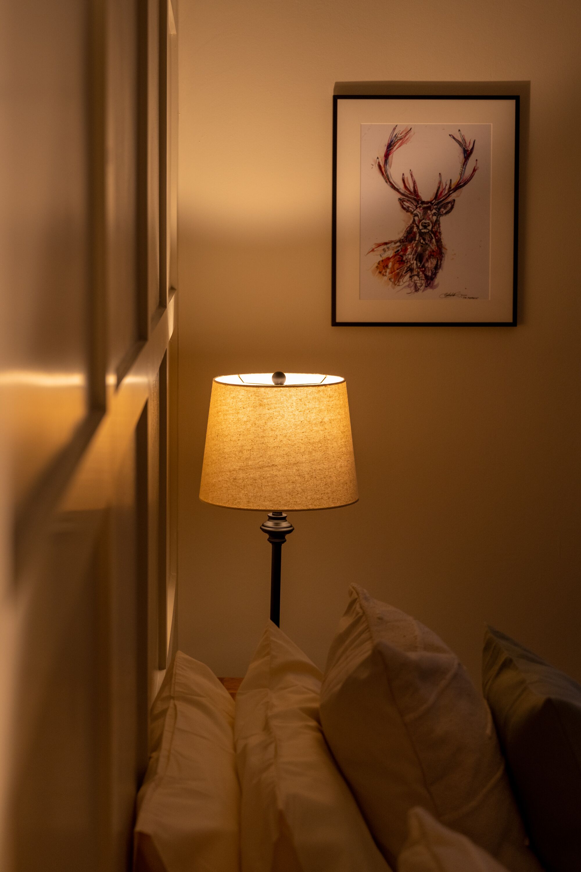 A room with a bedside lamp, a framed deer artwork on the wall, and a bed with pillows.