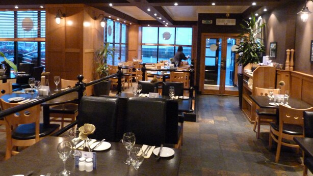Un restaurante con mesas dispuestas para comer, sillas de madera y vistas de la ciudad a través de amplios ventanales.
