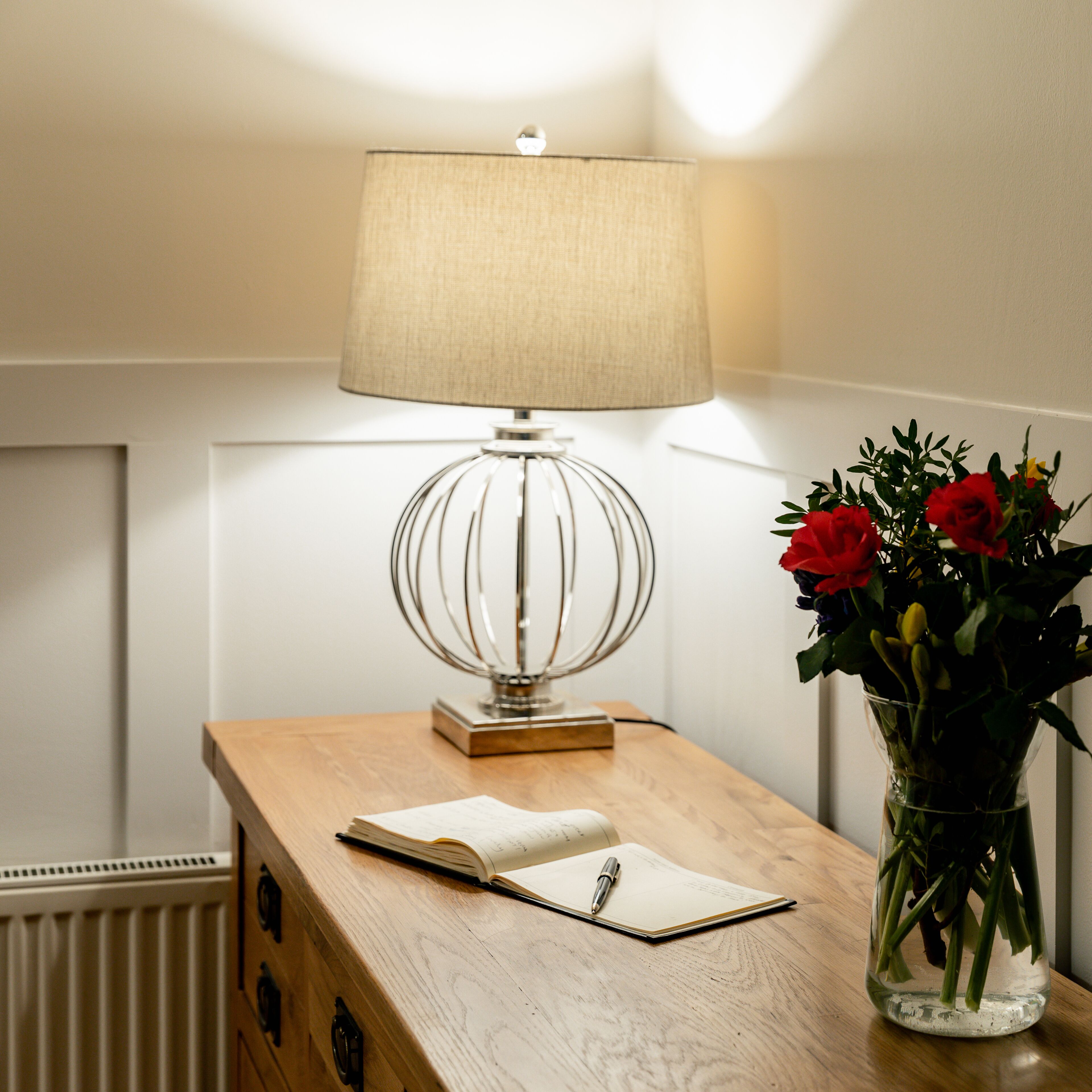 A wooden desk with a lamp, an open notebook, and a vase with flowers.
