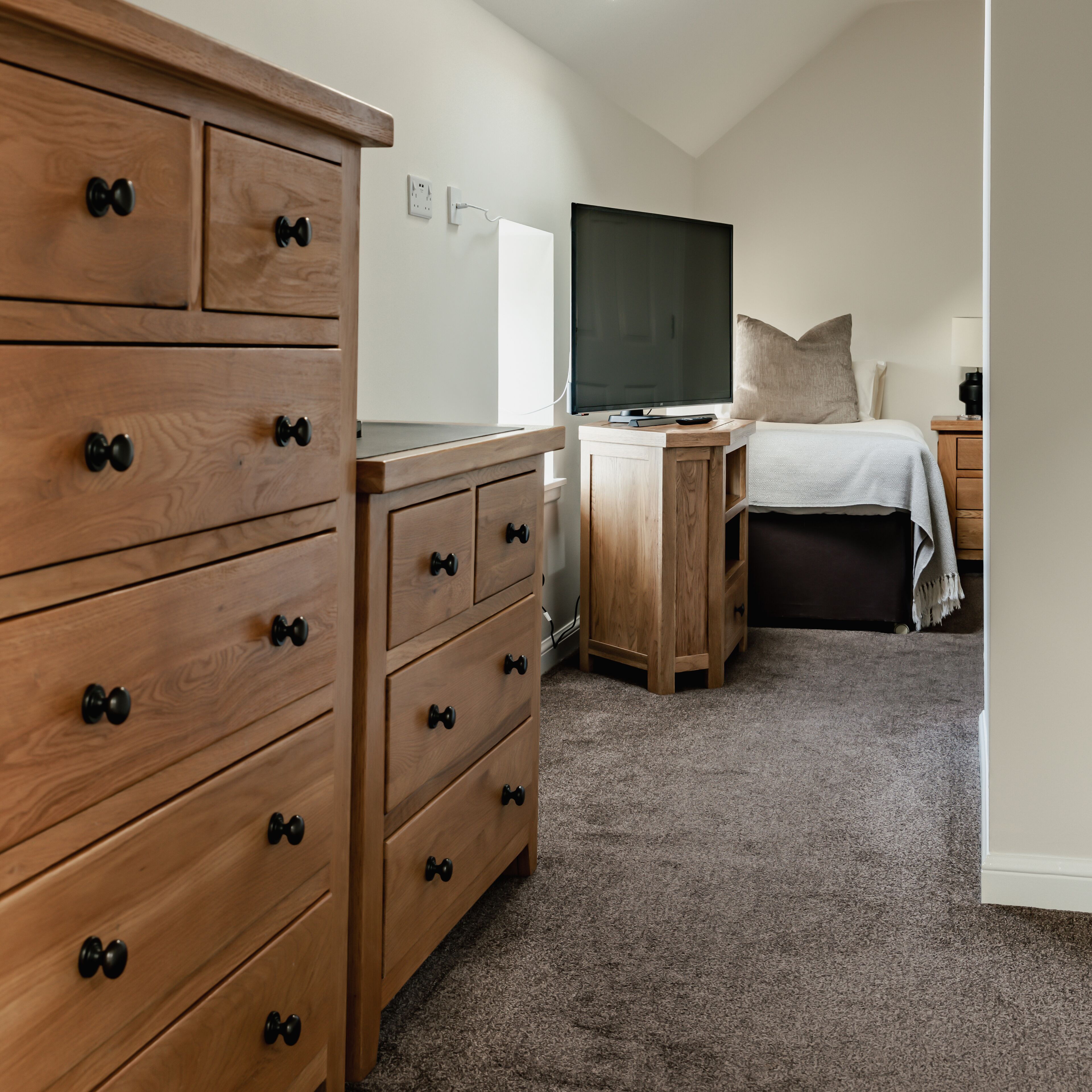Une chambre avec une commode en bois, un téléviseur posé sur un meuble, un lit recouvert d’une couette blanche et une table de chevet avec une lampe.