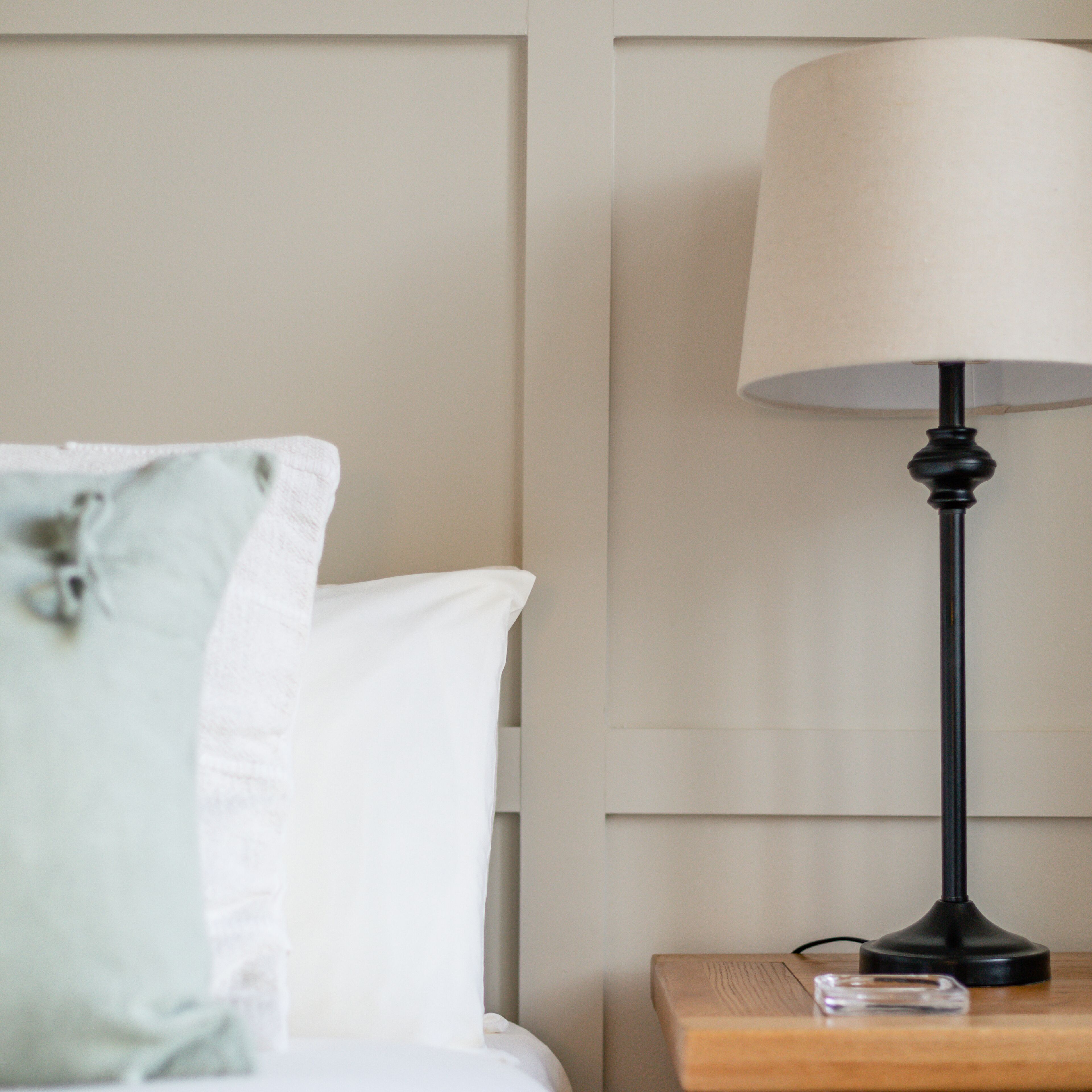 A neatly made bed with white and light blue pillows, a black lamp on a wooden nightstand, and a clear glass object on the nightstand.