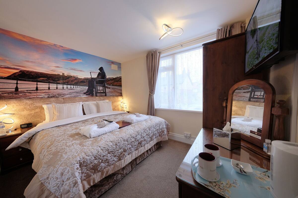 A hotel room with a large bed, a desk with a mirror, a TV, and a scenic mural on the wall.