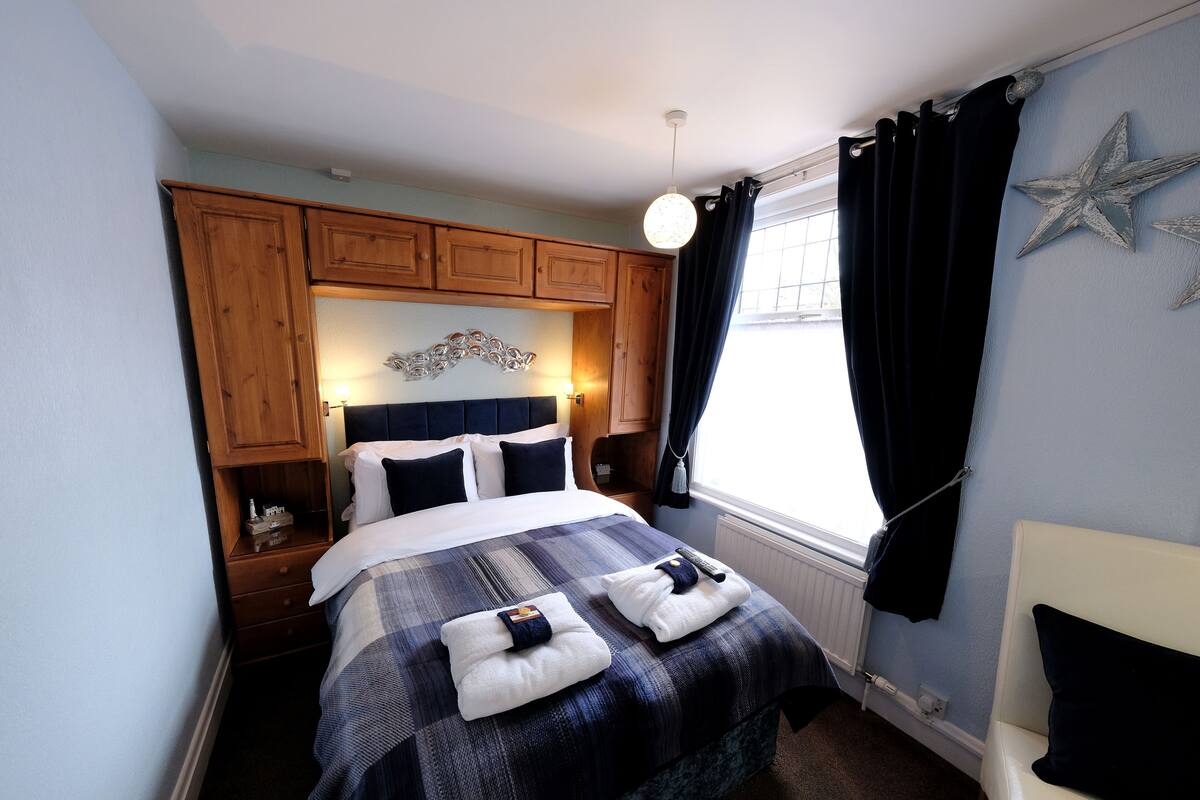 A neatly made bed with a blue and white checkered bedspread, two pillows, and two folded towels on the bed. There is a wooden headboard, a bedside table with a lamp, and a window with black curtains.