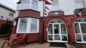 A two-story building with a red brick facade, a white-framed window, and a sign for Toulson Court. There are wooden chairs outside and a small Christmas tree. - Toulson Court (Scarborough)
