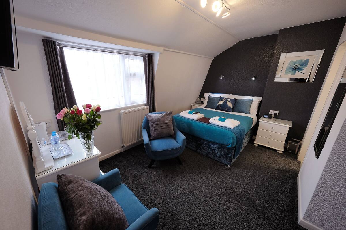 A compact hotel room with a bed, a desk with a chair, a small table with a vase of flowers, and a window with curtains.
