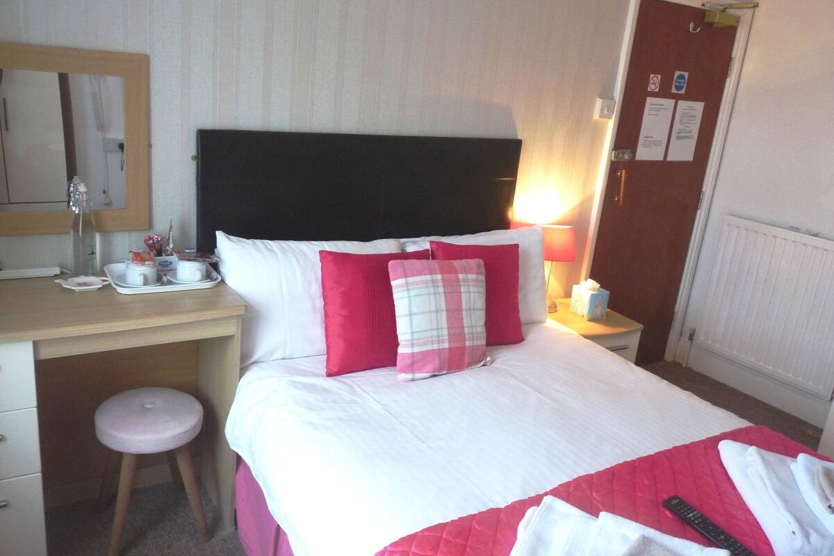 Quarto de hotel com uma cama, uma escrivaninha, uma cadeira, uma mesa de cabeceira e um espelho.