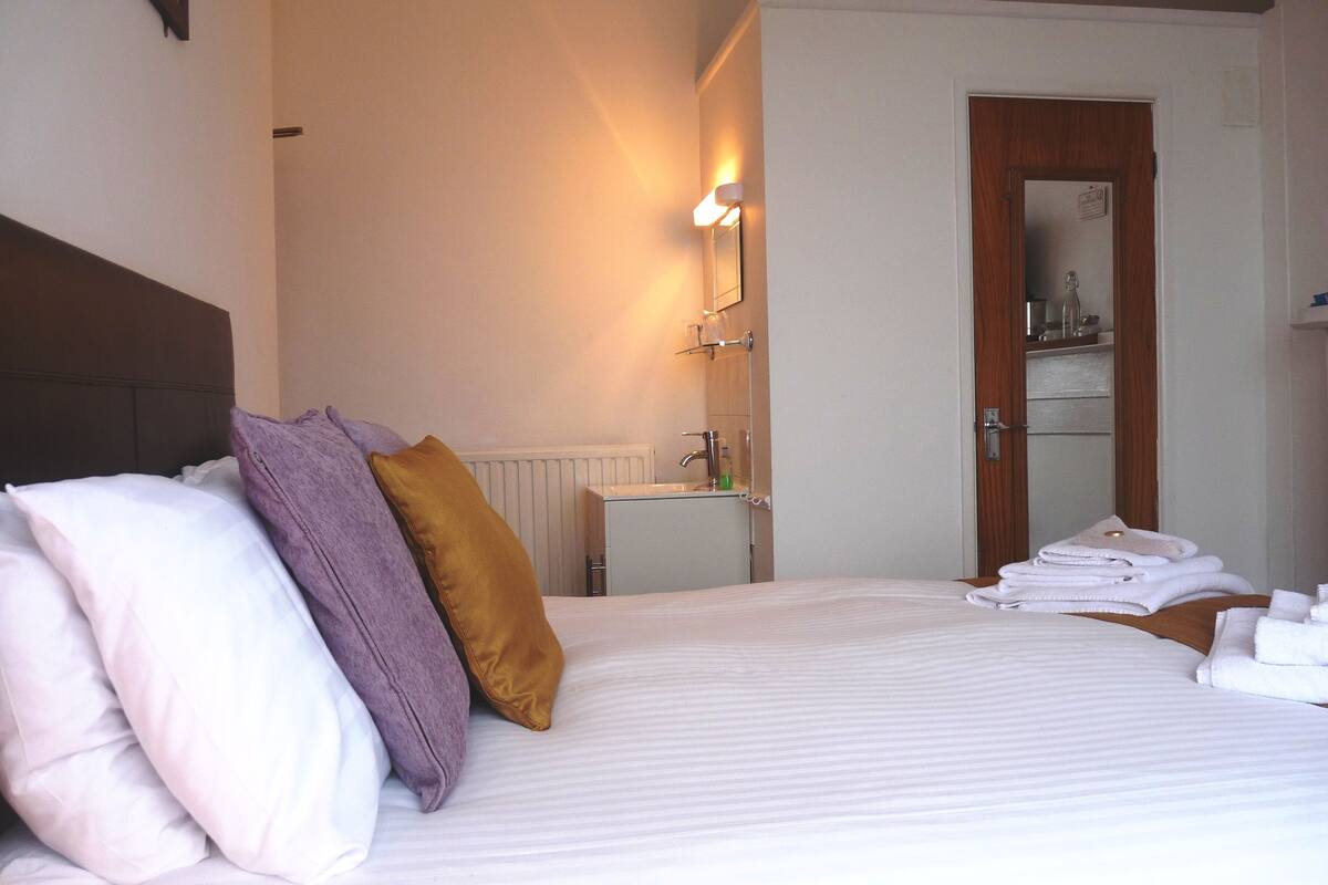 Quarto de hotel com cama, almofadas, mesa de cabeceira com toalhas, banheiro pequeno com espelho e porta que dá para outro cômodo.