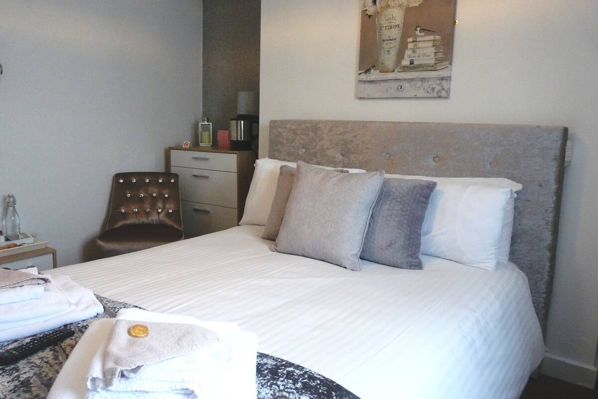 Uma cama bem arrumada com cabeceira, uma cadeira, uma mesa de cabeceira com abajur e um quadro pendurado na parede.