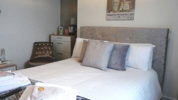 A neatly made bed with a headboard, a chair, a nightstand with a lamp, and a wall-mounted picture.