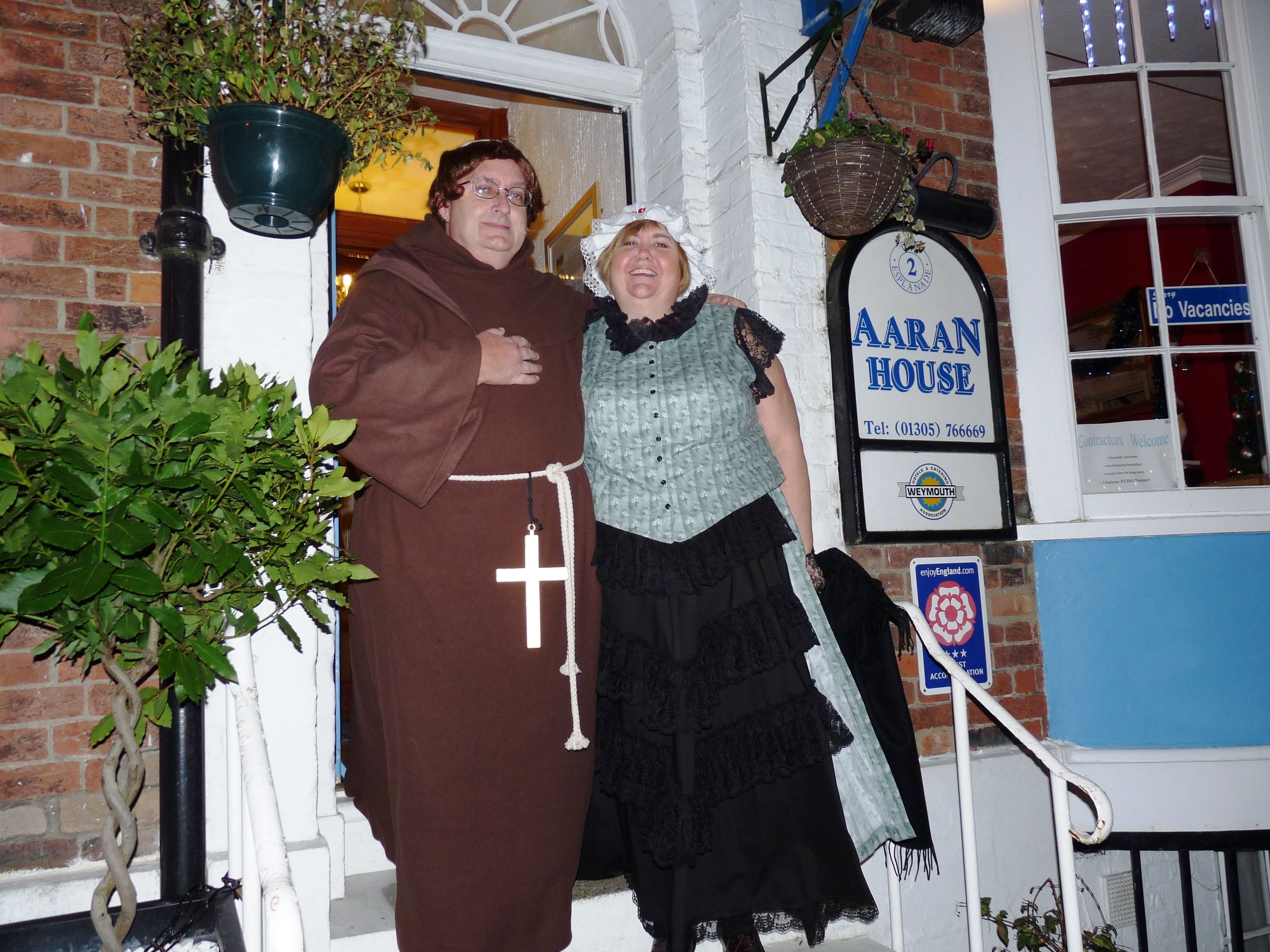 A person in a brown robe with a cross, standing next to a person in a blue and black outfit in front of a building with a sign reading "AARAN HOUSE".