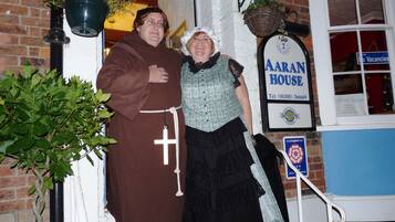A person in a brown robe with a cross, standing next to a person in a blue and black outfit in front of a building with a sign reading "AARAN HOUSE".