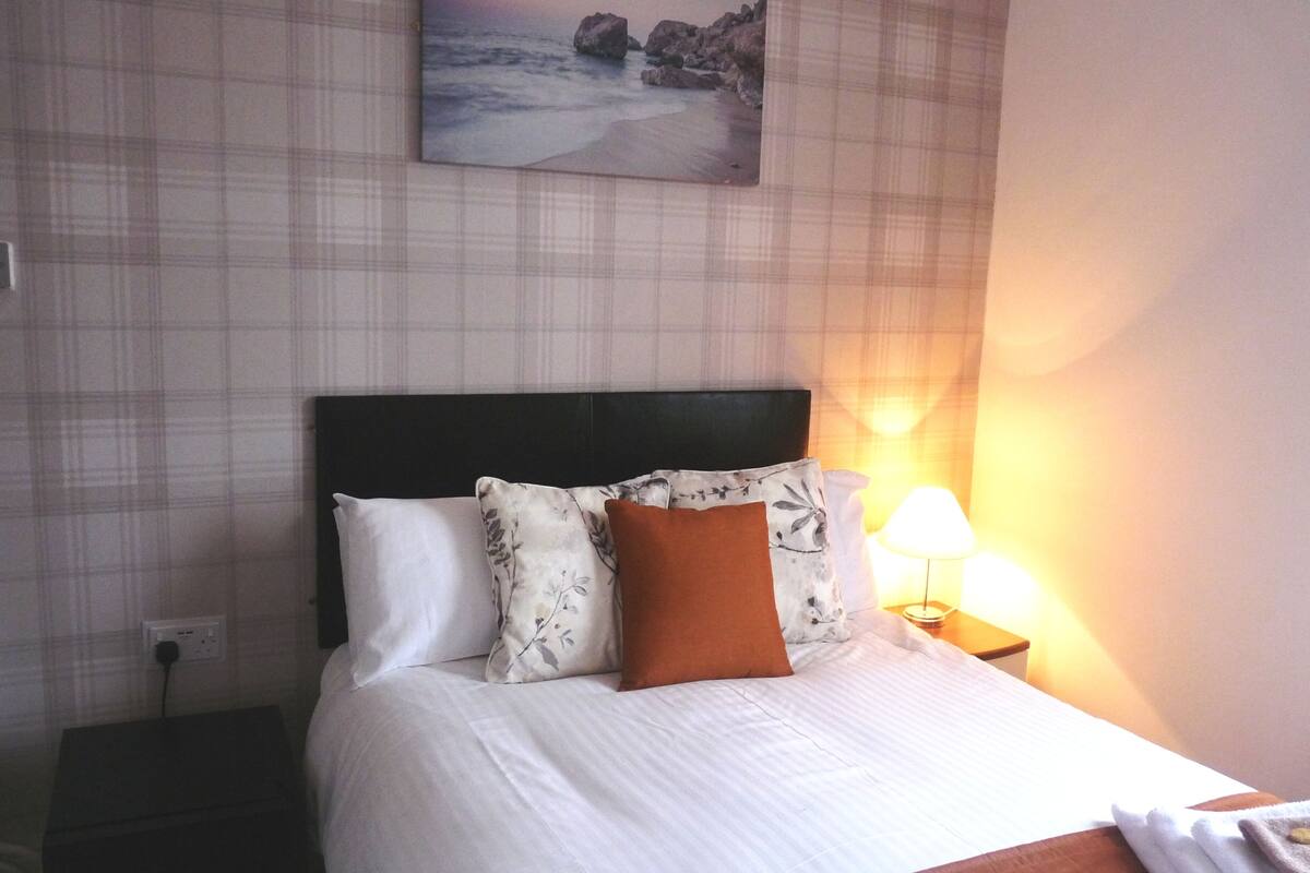 Quarto de hotel com uma cama, uma mesa de cabeceira com abajur e um quadro na parede.