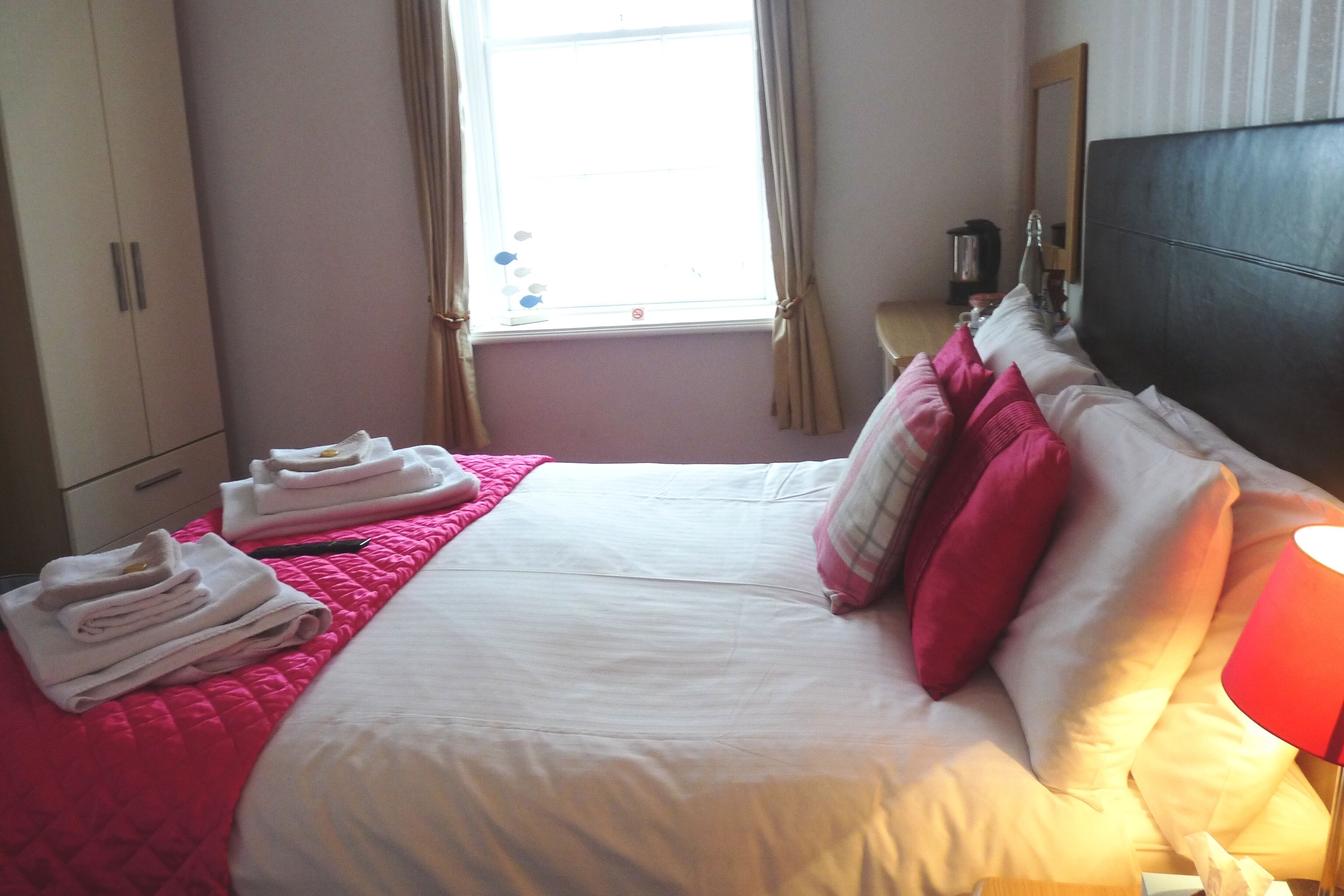 Habitación de hotel con cama, almohadas, mesita de noche con lámpara, ventana con cortinas y armario.
