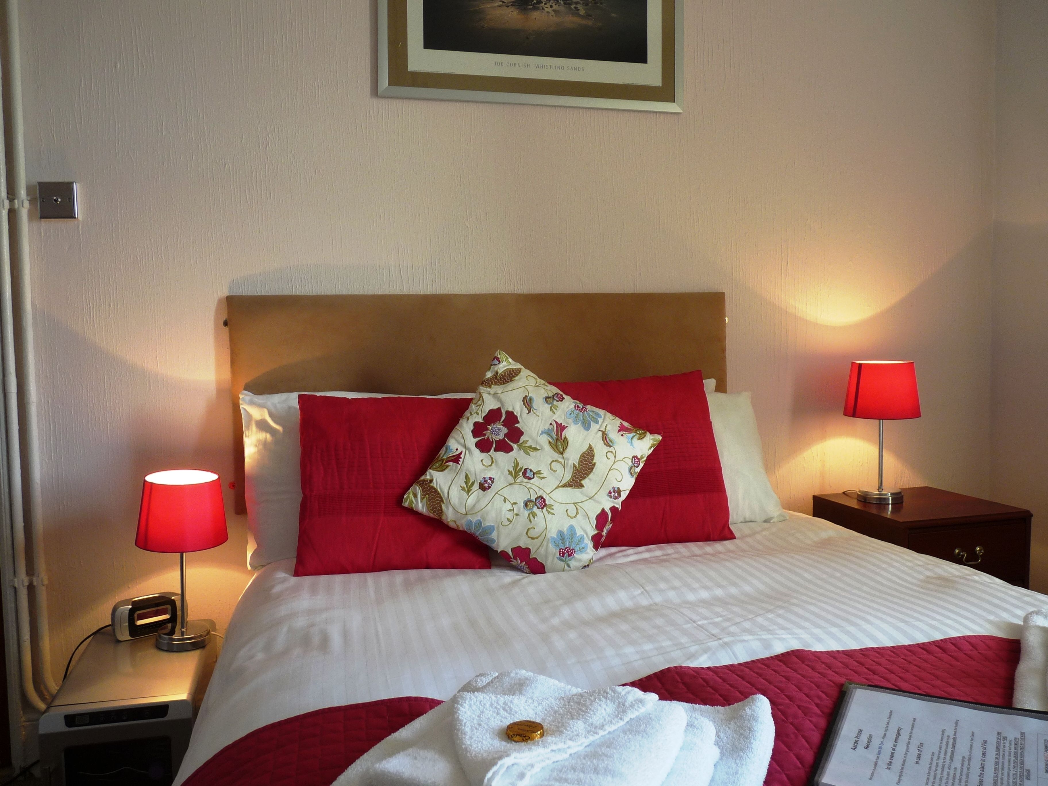 A neatly made bed with red and white bedding, two bedside lamps, a framed picture on the wall, and a sign on the nightstand.