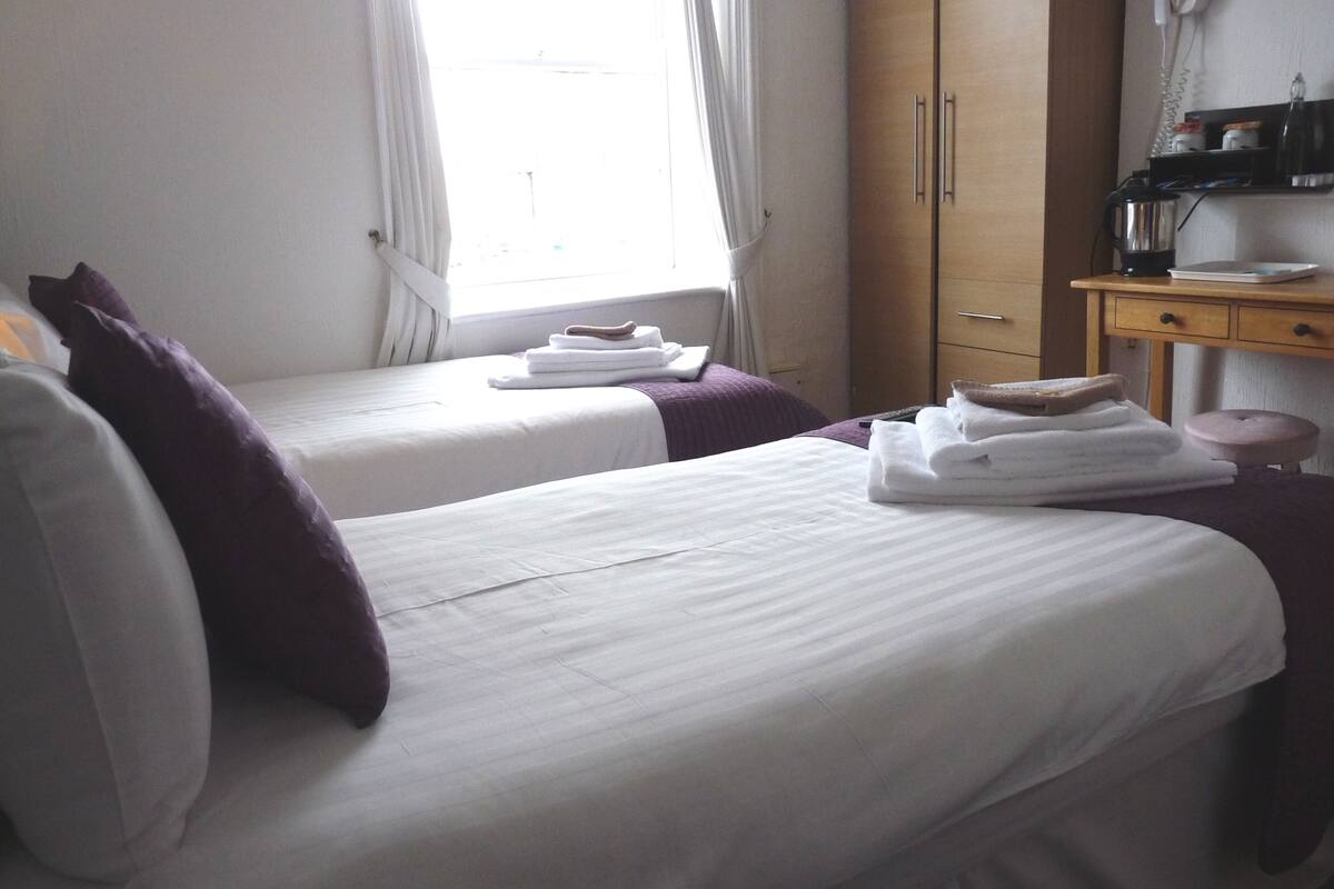 Quarto de hotel com duas camas, cada uma com travesseiro roxo e roupa de cama branca. Há um guarda-roupa de madeira e uma pequena mesa com uma chaleira e um prato.