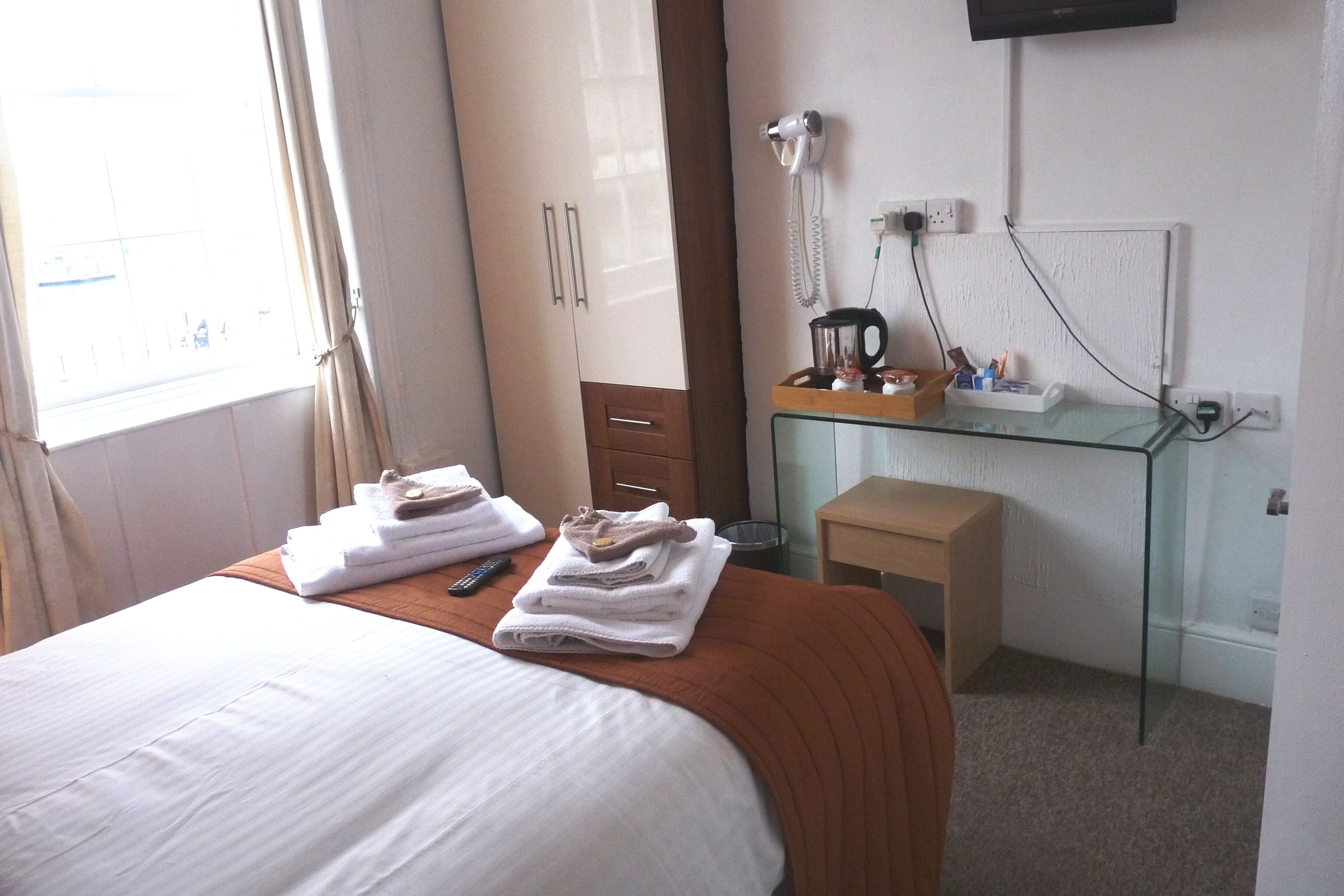 A hotel room with a bed, a glass desk with a coffee maker, a hairdryer, and a small wooden stool.